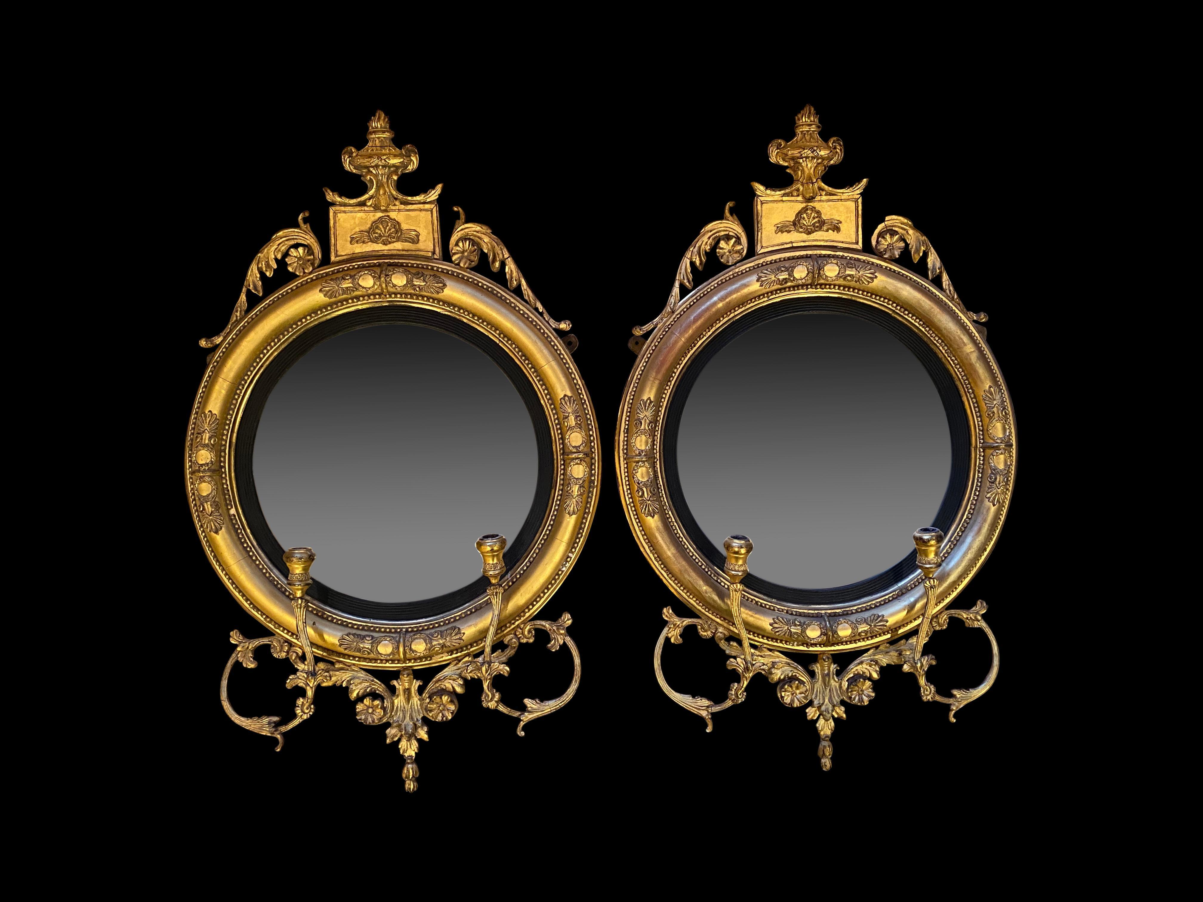 A fine pair of Regency carved giltwood and gesso convex mirrors, English, circa 1820. On top is a wonderful pediment urn. Each circular mirror plate with an ebonised and receded border, enclosed with a pair of gilded candle branches and foliate