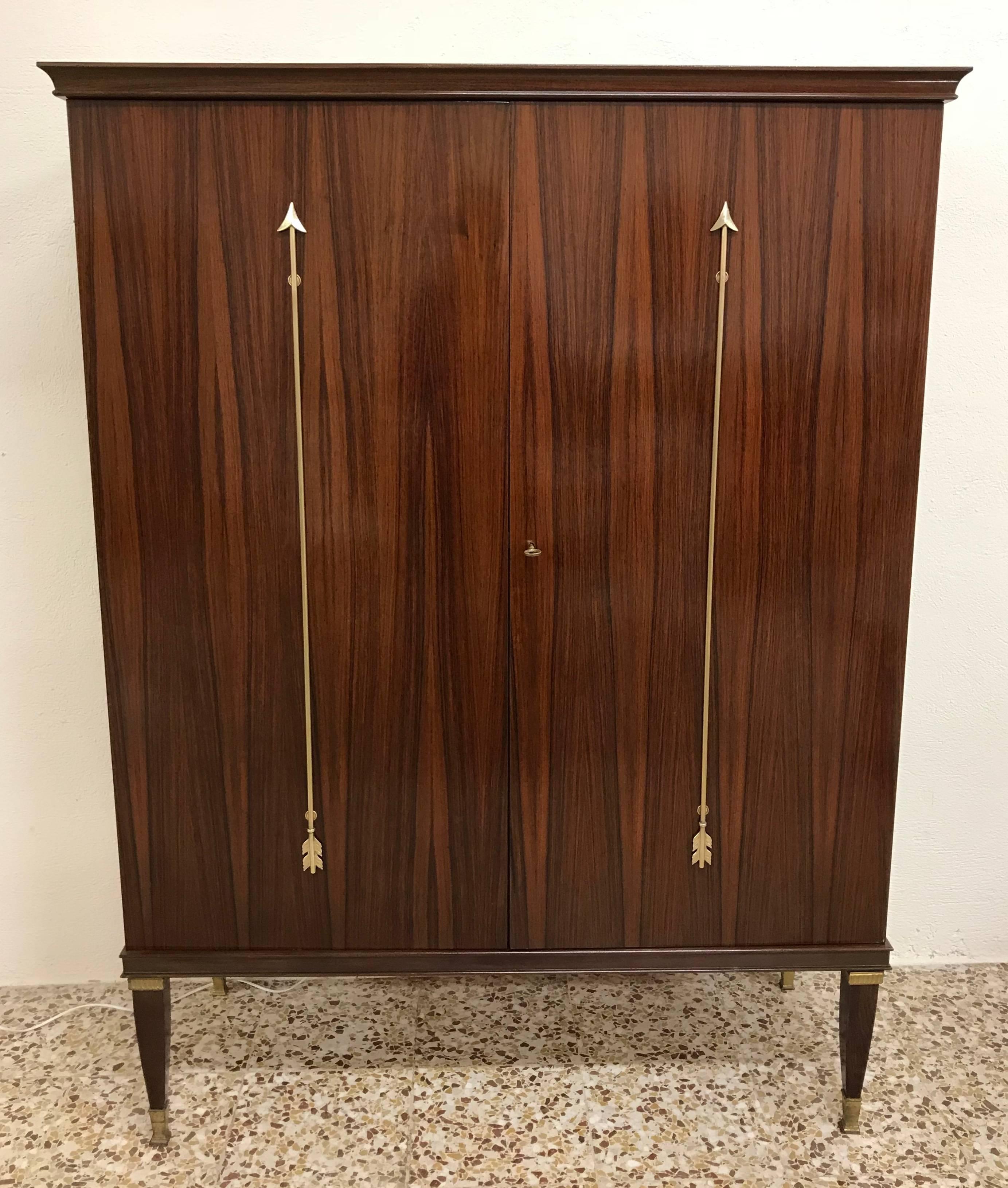 This large bar cabinet was produced in Italy in the 1950s by Paolo Buffa.
The central doors are embellished with two brass arrows.
The interior are finely fitted with drawers and cubbies, which are illuminated by lights switched on by doors opening.