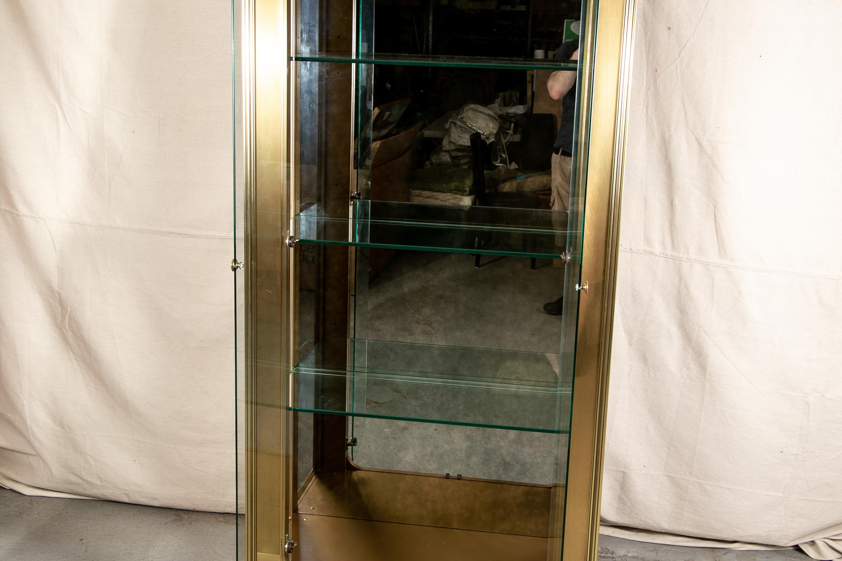 Fine tall brass display cabinet, reeded front frame with shaped top with palmette and scroll motif above the curved double glass doors with knob pulls. With glass side panels and three glass shelves. The lower frame with rosette and scroll motifs.