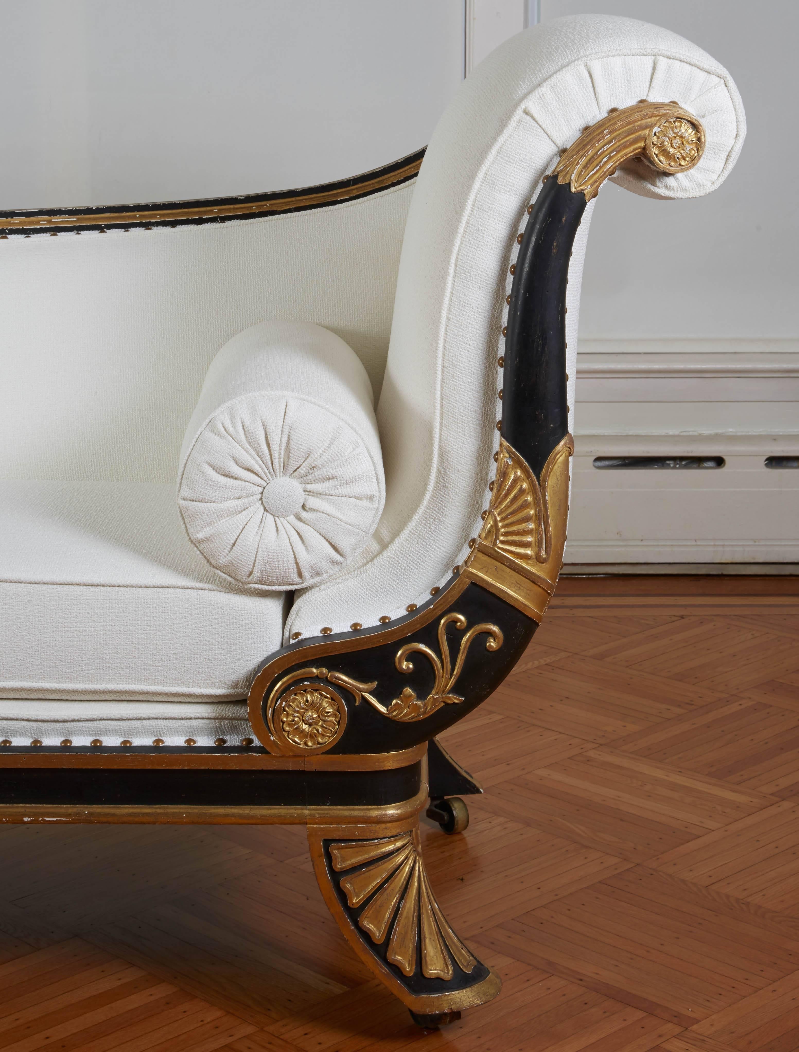 This daybed stands out for its impressive scale. With outward scrolling upholstered side and downswept back within an ebonized surround with gilt acanthus and lotus leaf decoration; the seat over a gilt-banded rail supported on outswept feet with