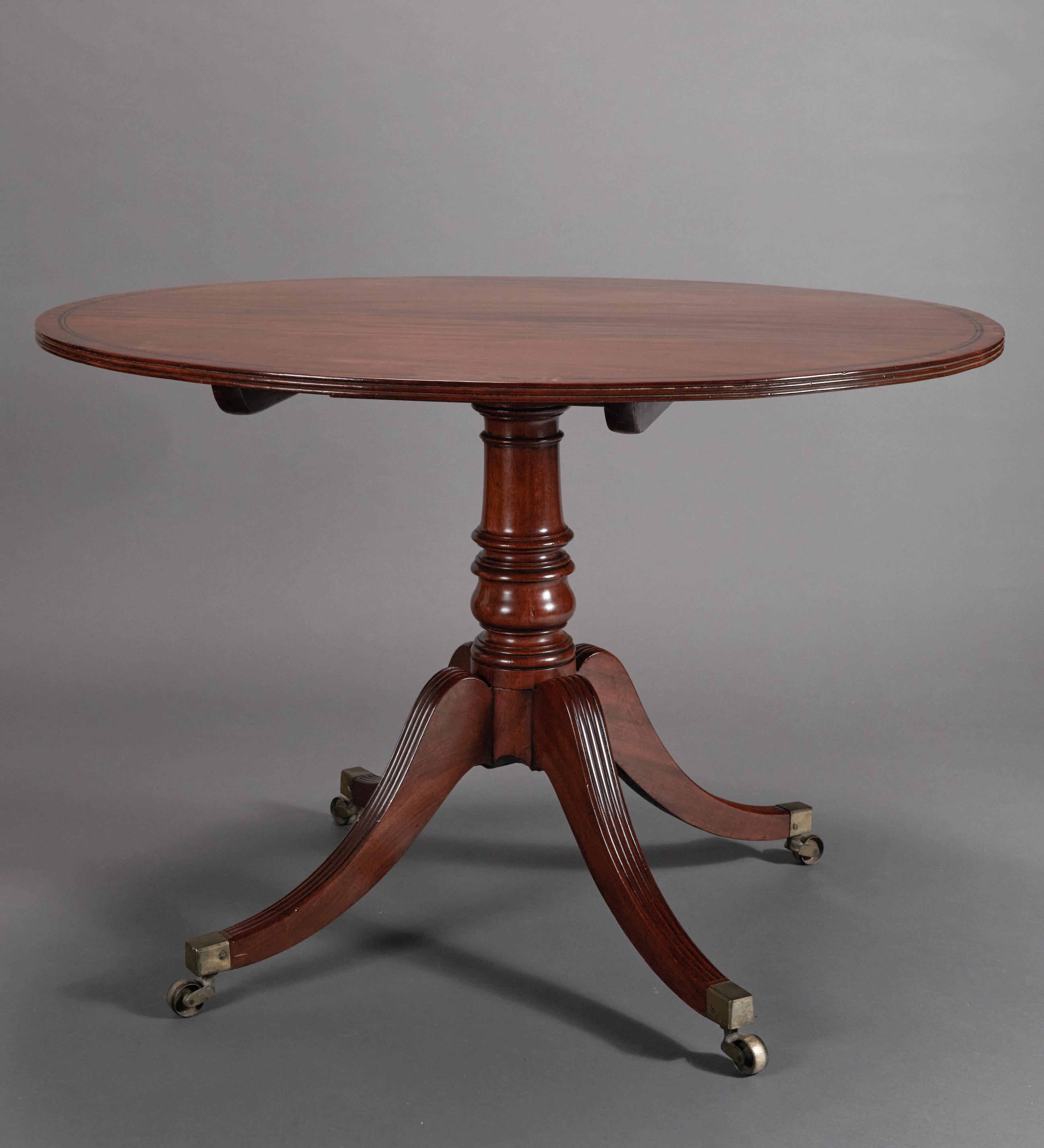 The circular solid mahogany tilt-top with two bands of ebony inlay, sits upon a turned pedestal with downward swept reeded legs with castors.
