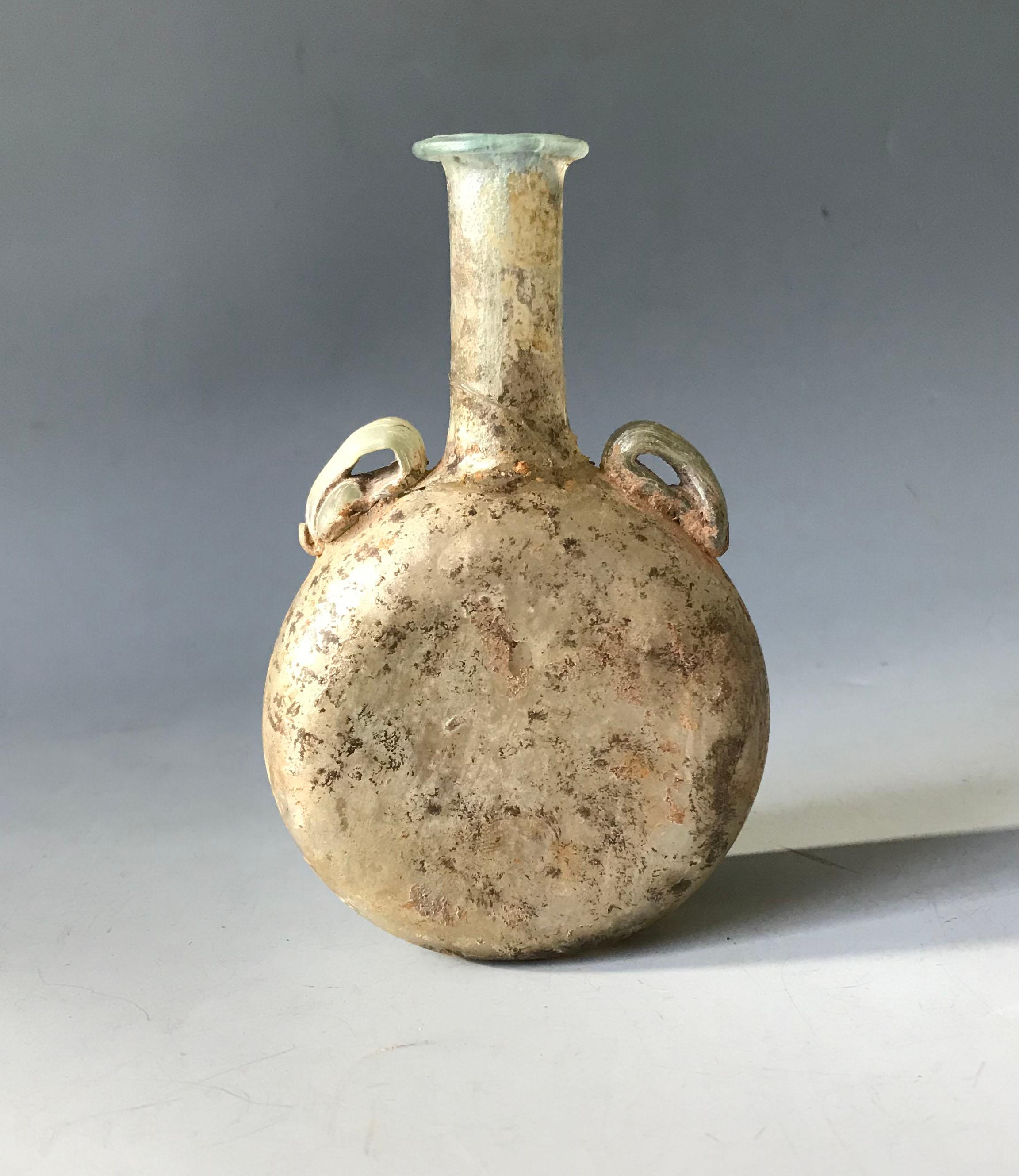 Roman iridescent glass flask antiquities ancient world.

A ancient Roman glass flask with round flattened body with thin neck and  2 applied handles.

Glass blowing was invented sometime early in the first century  in the Roman Middle East, it