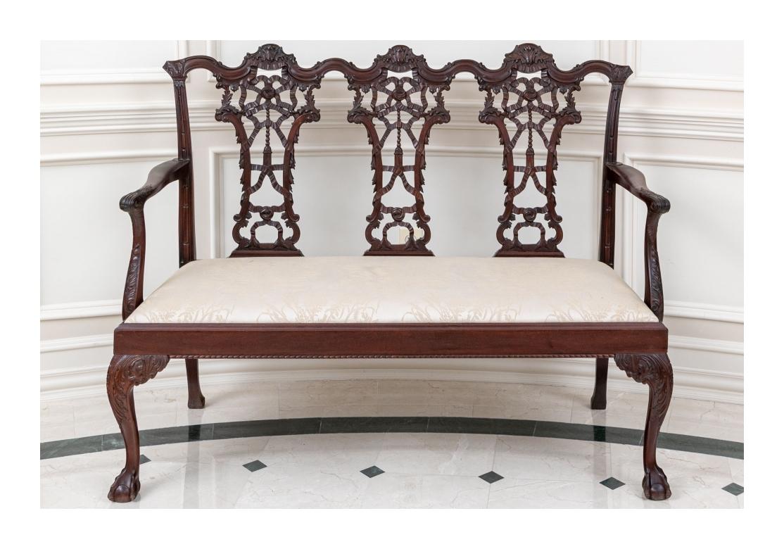 A intricately carved three seat settee with three part back in the classic Chippendale manner. The serpentine crest rail with scroll details and three openwork carved splats with ribbons, bows and leaves. The curved arms with supports decorated with