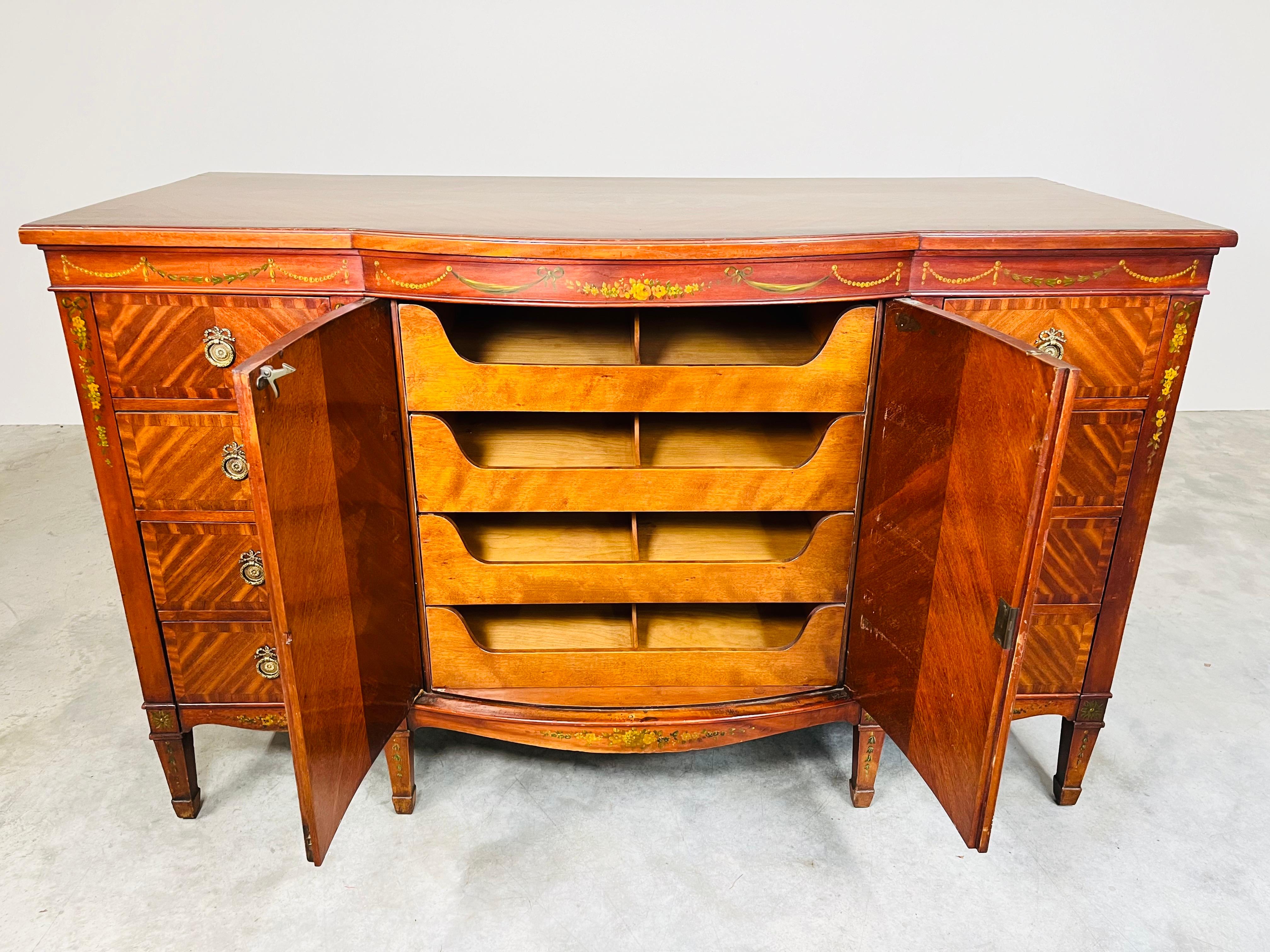 A fine hand painted satinwood veneer Sheraton Revival sideboard buffet having a frieze and bow front pair of doors painted in Adams style with neoclassical swags, paterae with scrolls and bellflower pendants, having 4 pull out interior drawers or