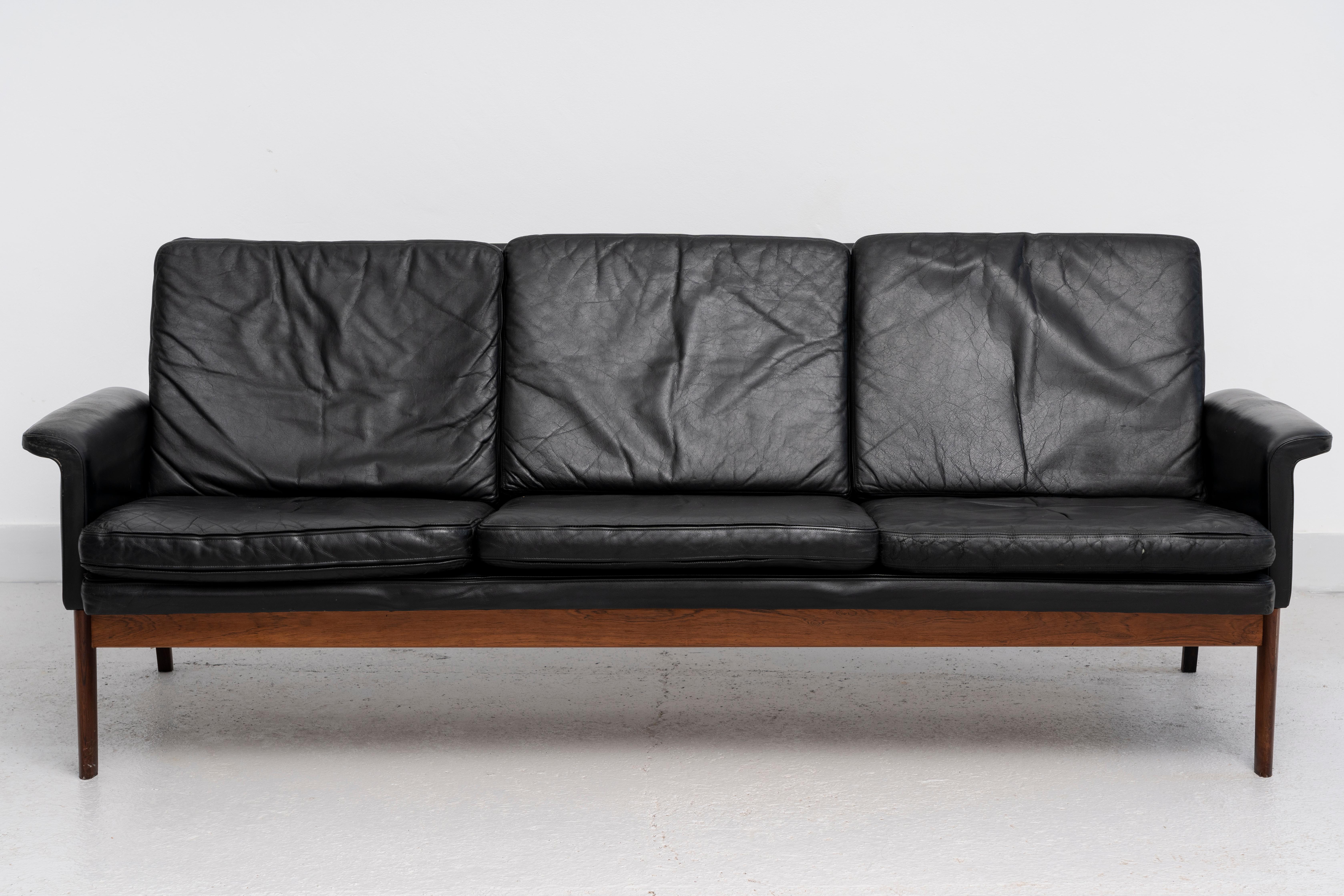 Sofá Júpiter, modelo 218 de cuero negro y madera de frutal. Con etiqueta y número de serie 6635134. Tiene desgaste en el cuero y algunos desgarros. Este modelo fue diseñado por Finn Juhl y fabricado por France and Son en 1965. El sofá tiene un