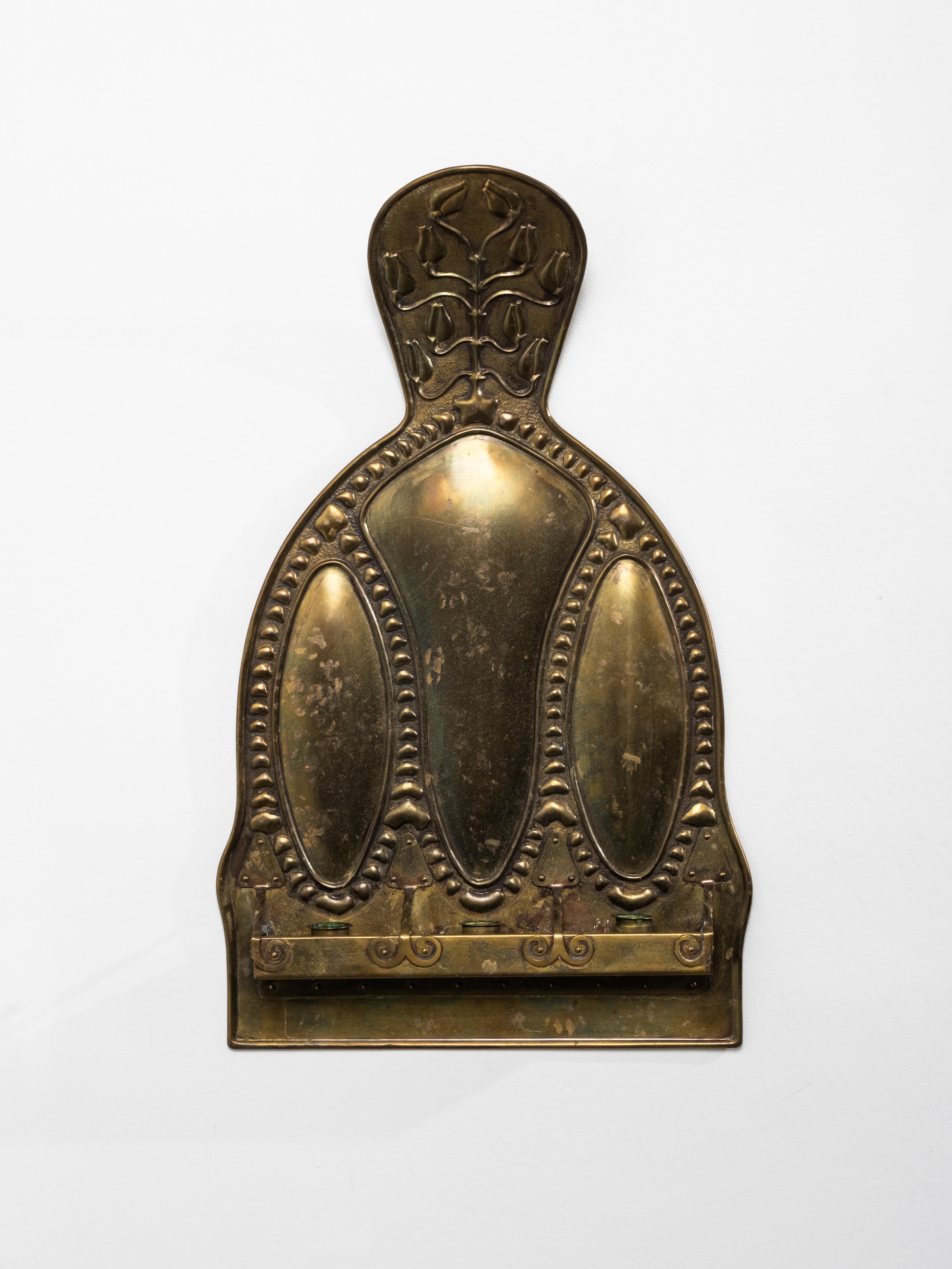 Este aplique de latón de principios del siglo XX tiene una forma de marcada influencia Art Nouveau, caracterizada por curvas suaves, ornamentación en relieve y motivos naturales. Los tres paneles ovalados alargados crean un ritmo vertical