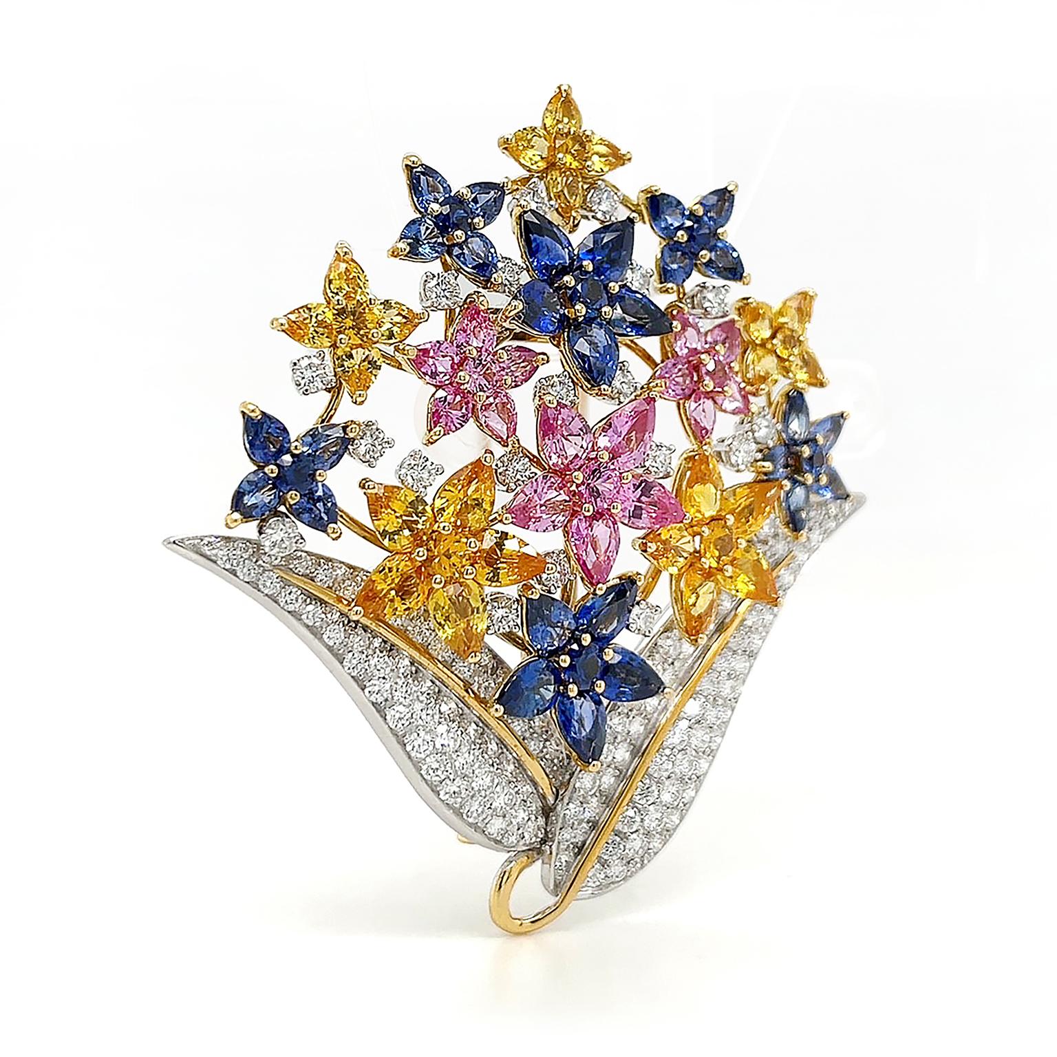 Bei dieser Brosche schimmern Saphire in vielen Farben als Edelsteinblüten. Aus zwei schlanken Platinblättern, die mit Diamanten im Brillantschliff und Goldakzenten verziert sind, sprießen Gelb-, Rosa- und Blautöne. Birnenförmige Saphire umgeben