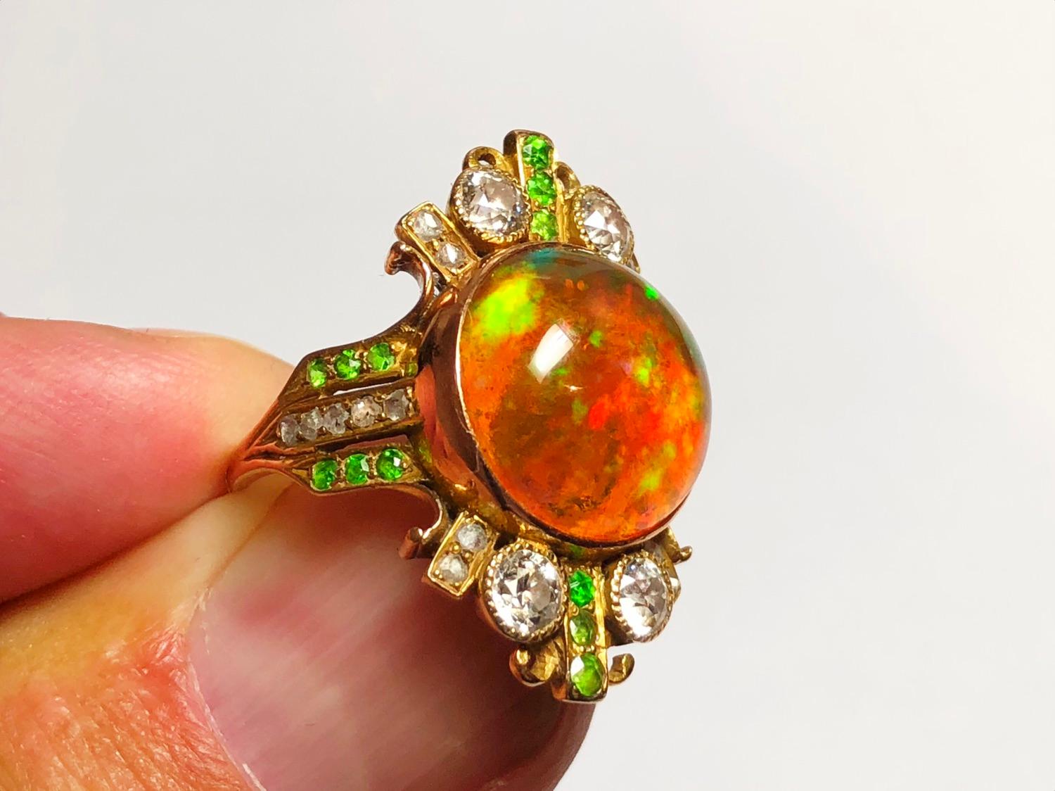 A fire opal and demantoid garnet ring, set with a central fire opal, with play of colour, in a decorative gold mount, with surrounding demantoid garnets and old-cut and rose-cut diamonds.

Opal diameter 13mm. Finger size O / USA 7