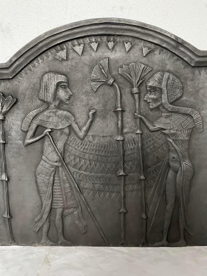 The design of this modern fireplace plaque draws inspiration from the art of ancient Egypt.

On the left, a man wearing an usekh collar and dressed in a shendyt (the traditional Egyptian loincloth) leans on a staff and converses with a woman facing