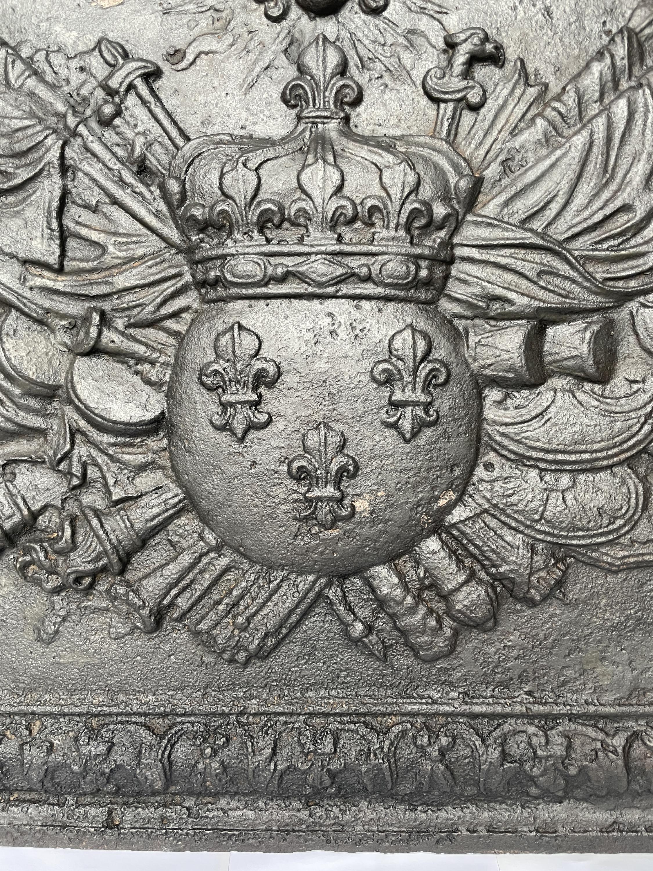This exceptional fireback was cast in the 17th century.
It is adorned by the France's coat of arms crowned with the Royal crown put in front of flags and war weapons. We can also see the mascarons depicting the Sun King and his motto 