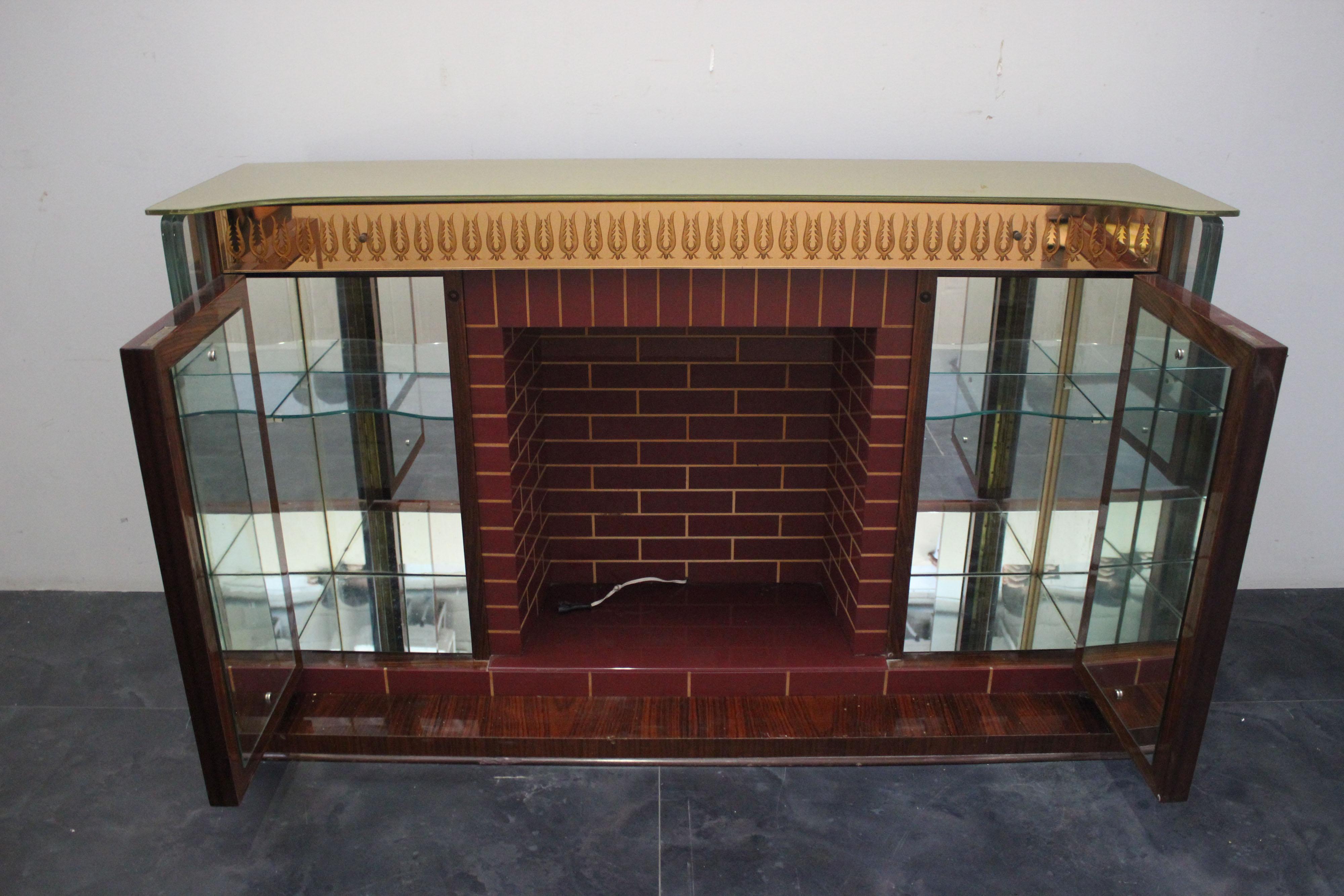 Singular bar cabinet in the form of a fireplace. Rosewood sides and base. On the front it has burgundy glass rectangles profiled in maple imitating masonry, doors and under-top band with mirrors engraved with classical motifs with gold appliqué.