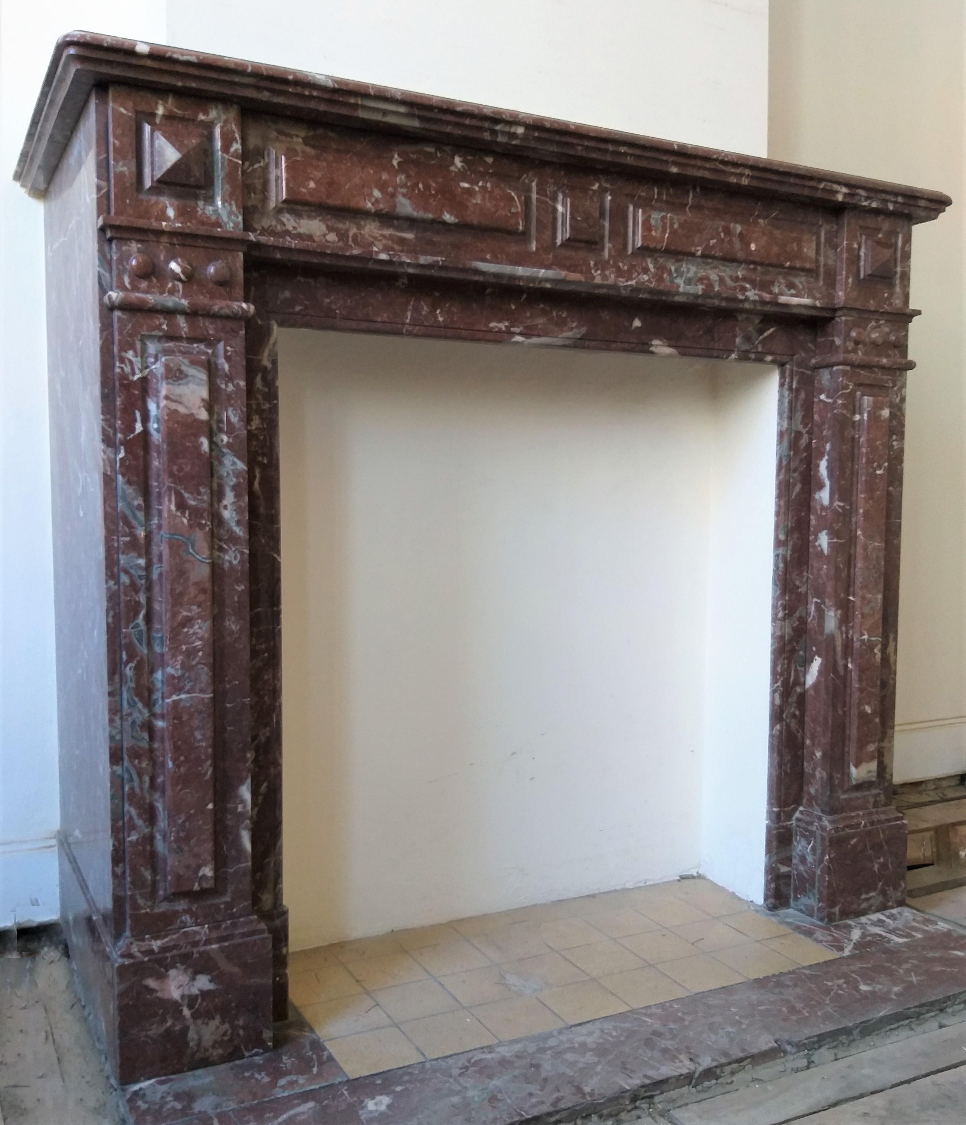 This is a late 19th century, polished Belgian Rance marble,  fireplace-surround.  The moulded shelf rests above a paneled frieze with a central lozenge and cushioned marble panels.  Flanked by square bossed endblocks.  All is echoed on the jambs. 
