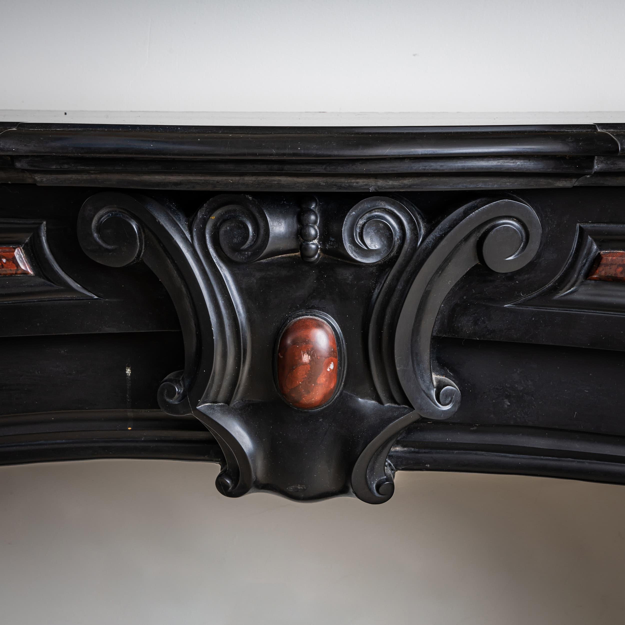 Grand, courtly fireplace surround crafted from black stone with refined inlays of Rouge Griotte, a richly veined deep red marble. The design is inspired by Renaissance motifs, featuring large volutes and a central cartouche.
Executed in heavy,
