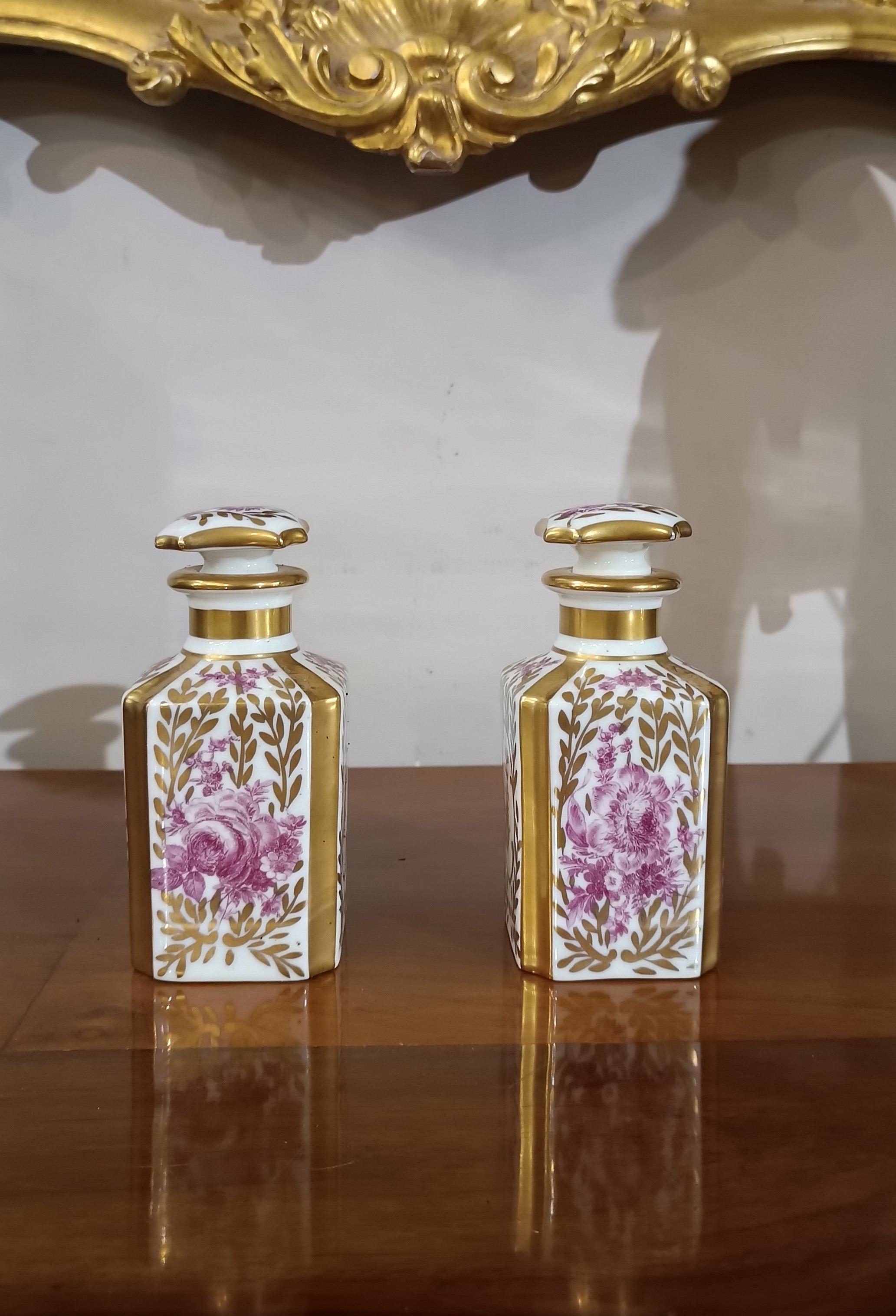 Elegant pair of small perfume bottles in white porcelain, decorated with gold details and painted in a refined burgundy color. On each side of the bottles and on the cap there are floral decorations depicting four different types of plants and