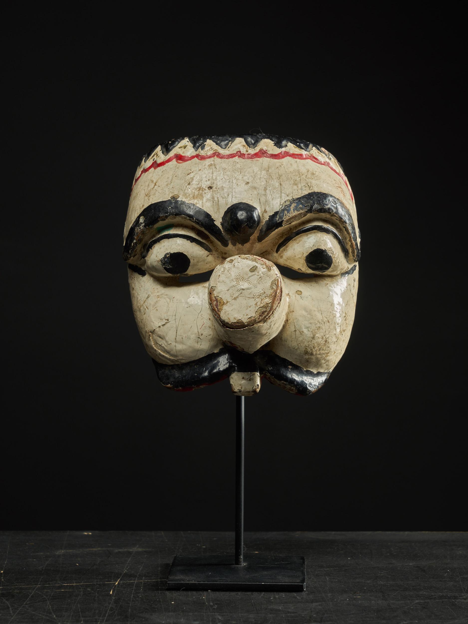 The mask is an example of traditional theater masks found in the cultures of Java, Indonesia. The face is carved out of wood and painted with colors that represent the status of the character. In this case, the mask is almost all white, which is a