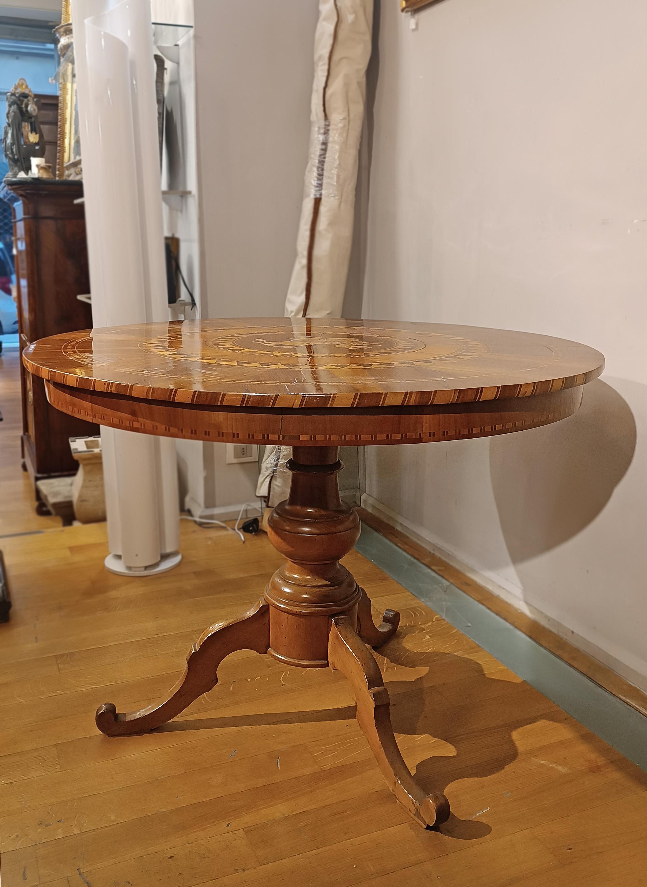 Elegante mesa Rolo en madera de nogal, finamente tallada y enriquecida con incrustaciones de madera de frutal. La estructura se caracteriza por una pata central moldeada y tres pies con volutas, que le confieren estabilidad y refinamiento. El