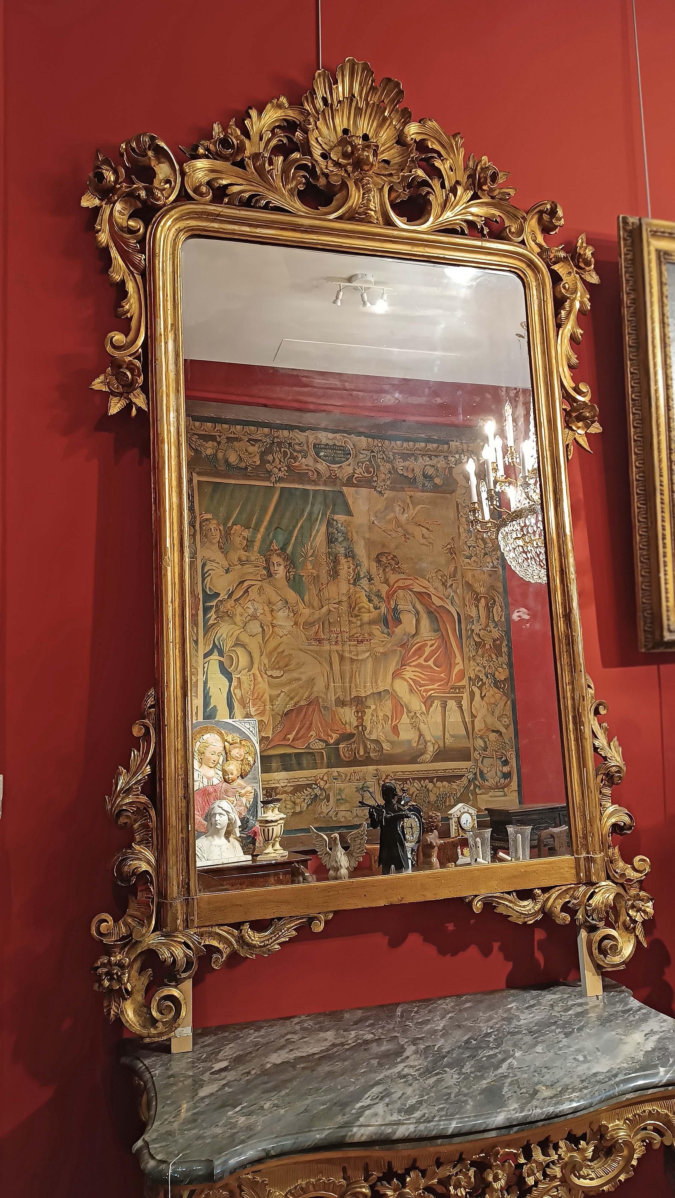 Elegante specchio di grandi dimensioni in pino svizzero, intagliato e dorato con foglia d'oro zecchino. La cornice è ornata da decorazioni nella parte superiore e inferiore, con grandi riccioli e intrecci vegetali che ne accentuano l'eleganza,