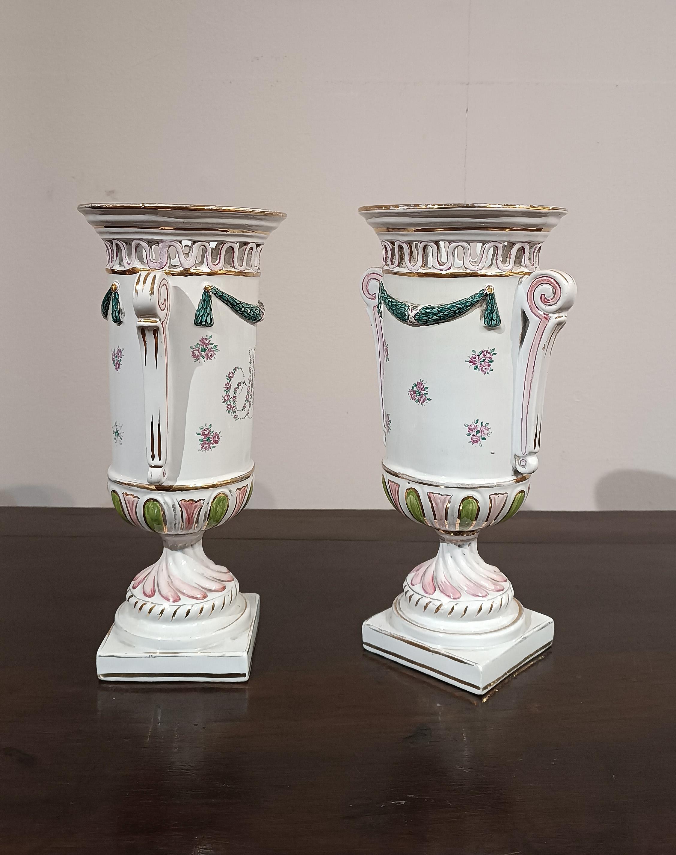 Una raffinata coppia di vasi in porcellana dipinta, impreziositi da finiture in oro. I vasi sono caratterizzati da una forma ad anfora, con una base quadrata e manici arricciati. Ad aumentare l'eleganza di questa coppia sono le decorazioni, con