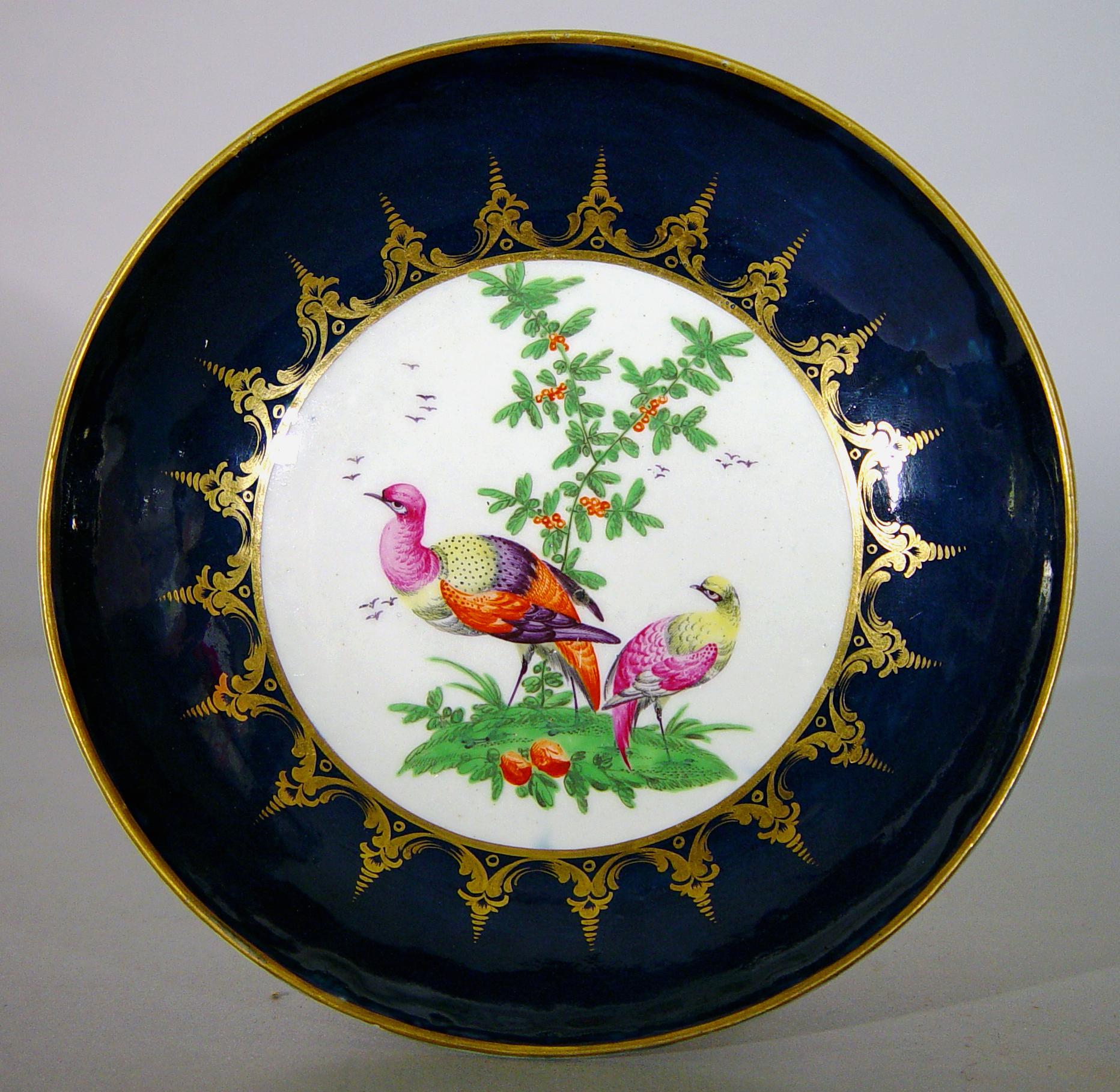 First Period Worcester Porcelain blue-ground exotic bird-decorated cake plate,
Circa 1770.

The First Period Worcester saucer dish is decorated with mazarine-blue ground. The central circular reserve is painted with two exotic birds standing on