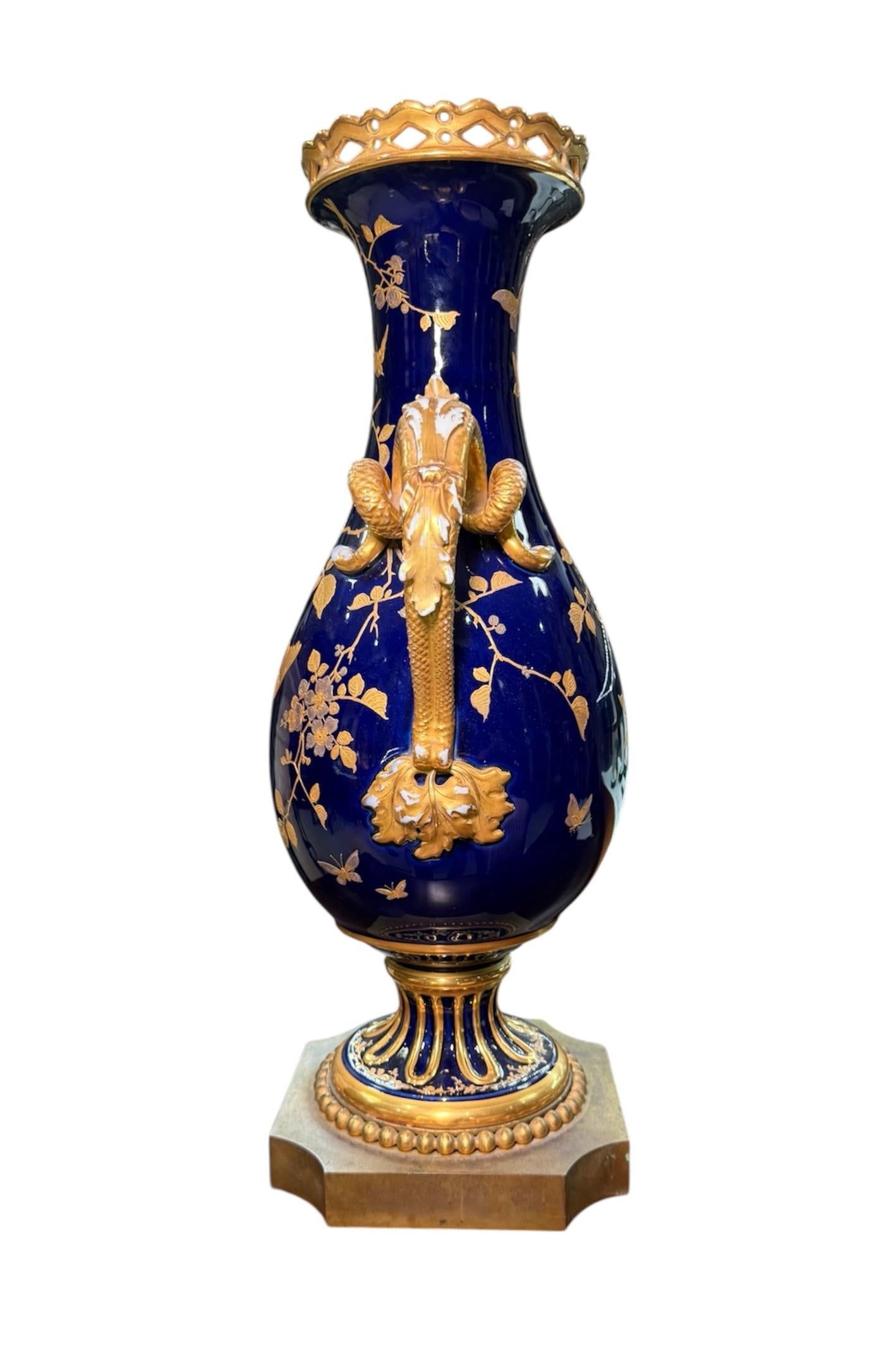 This is a Fischer and Mieg Porcelain Urn Vase. It depicts a tall cobalt blue urn decorated with long gilt branches of flowers, leaves and birds. Both gilt handles of the urn are shaped as snakes covered in the center by acanthus leaves. The upper
