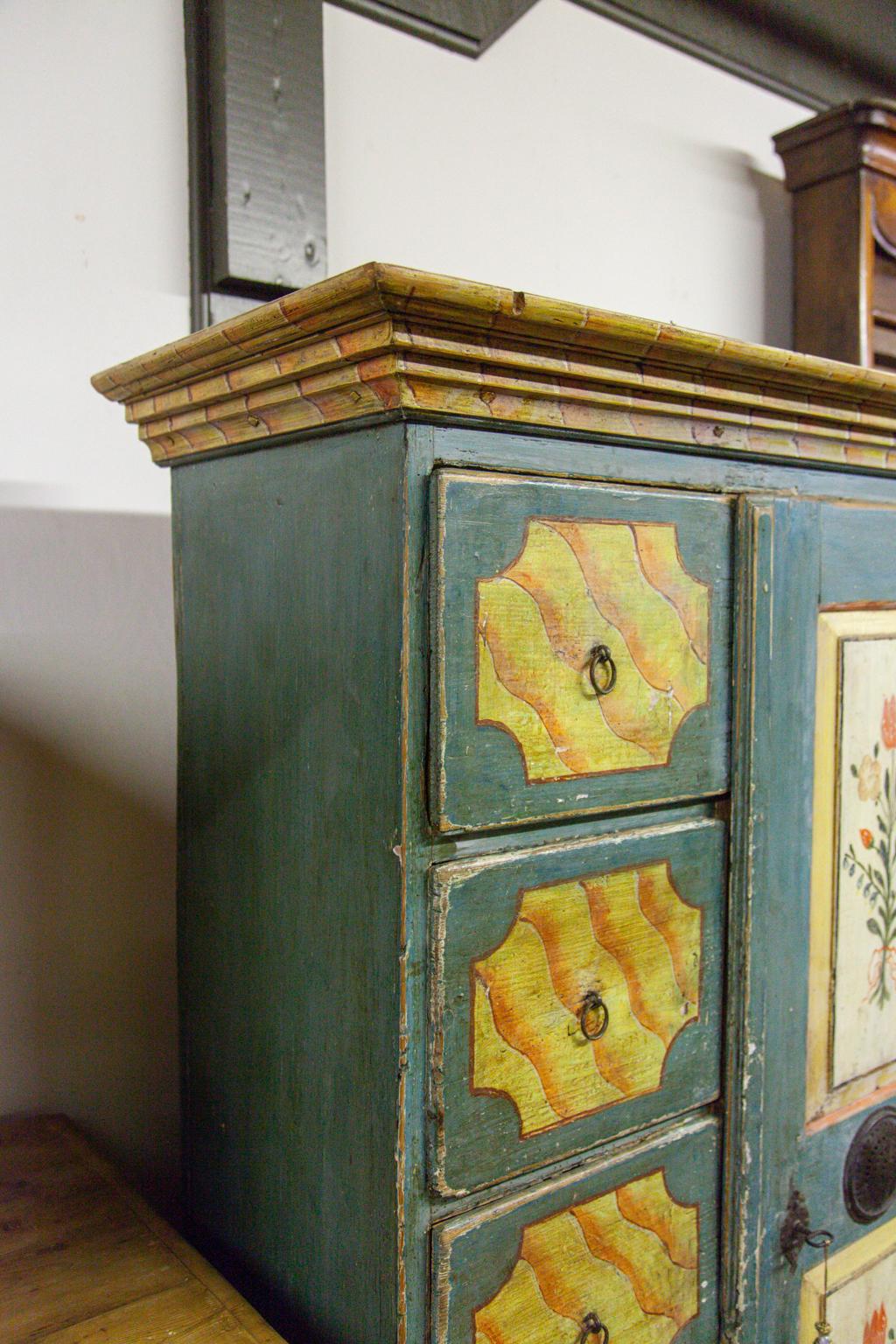 Five-drawer faux painted side by side cupboard, the door with raised floral painted panels and perforated circular tin vent in center, attached with exposed steel hinges; the five drawers with faux painted angular design that is repeated in the cove