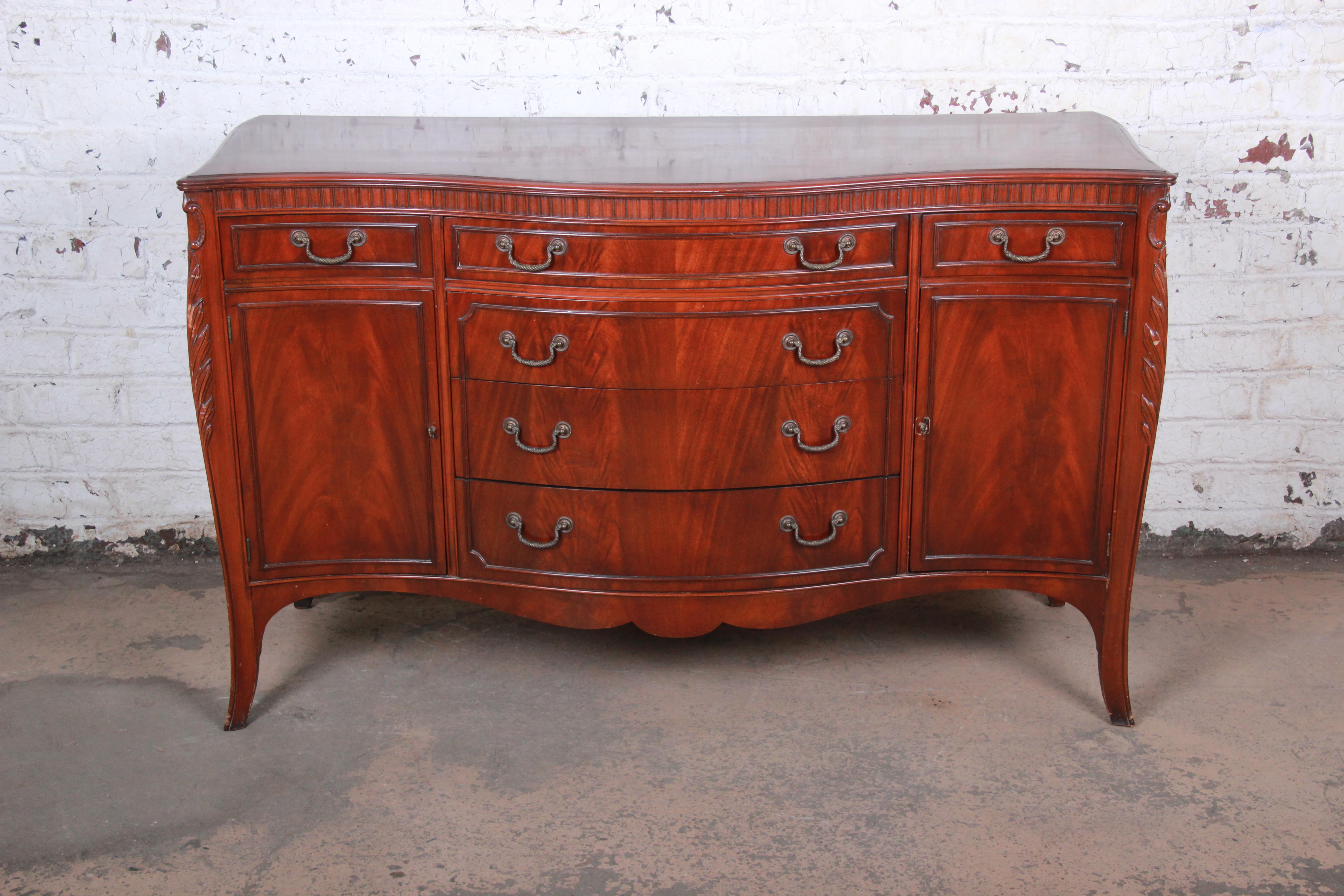 A gorgeous vintage bow front French carved sideboard or credenza by Drexel Furniture. The sideboard features stunning flame mahogany wood grain and nice carved wood details. It offers ample storage, with six dovetailed drawers and two shelved