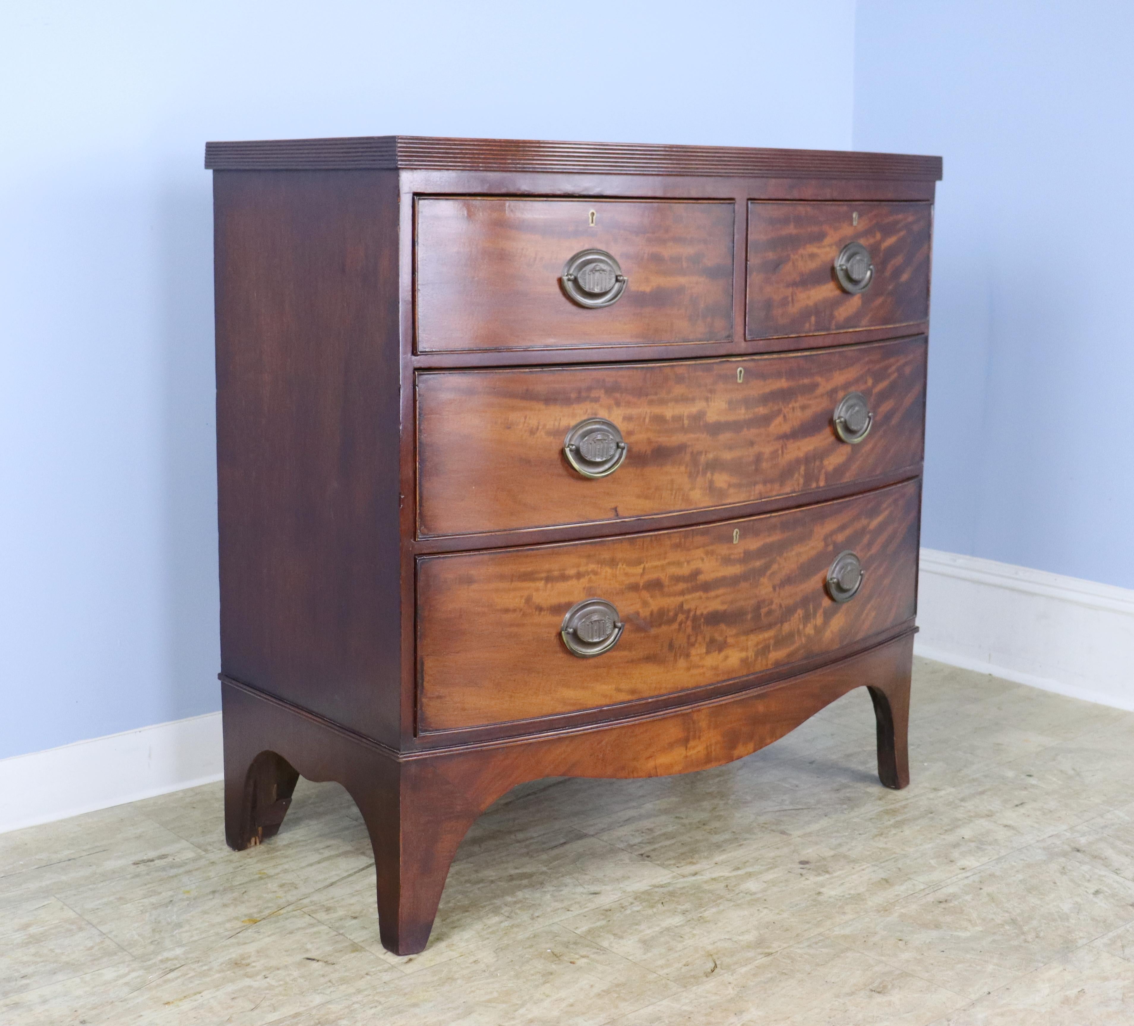 An antique bowfront bureau from England. Note the dramatic flame mahogany grain as well as the cockbeaded edging around the drawers, which are quite clean on the interior. Wonderful detailed reeded edge around the top. Original handles and feet.