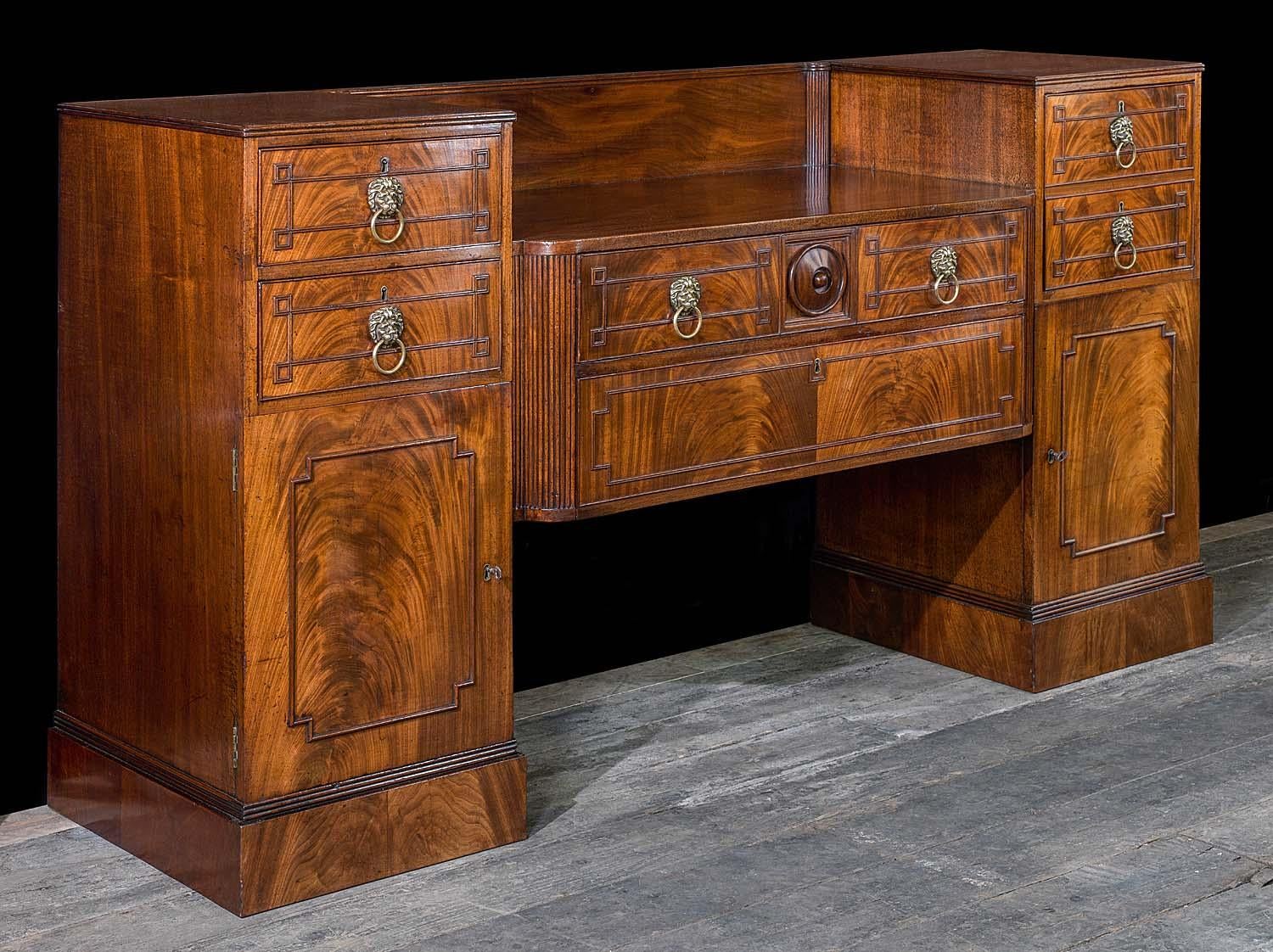 An early Regency twin pedestal sideboard in flame mahogany with a bowed tablet frieze which is flanked by two dovetailed drawers and a lower cupboard to the left, and a single dovetailed drawer mounted with a wine holder and lower cupboard to the
