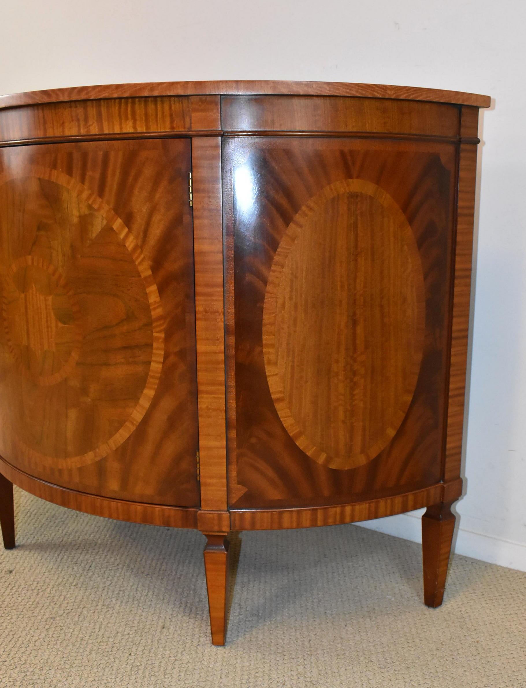 A beautiful flamed mahogany demilune by Baker Furniture as a part of their Fine Historic Charleston Collection. This piece is adorned with a mix of exotic wood inlays. The front is decorated with banded ovals surrounding a central starburst; the