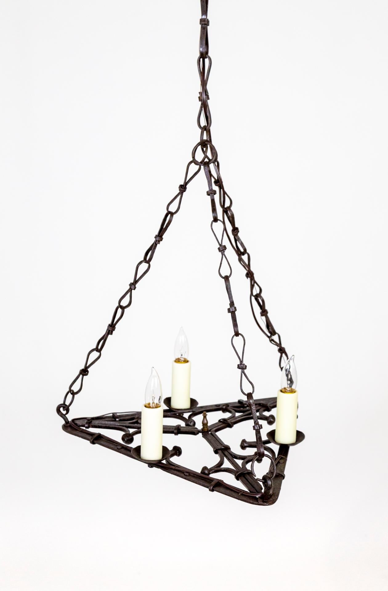 A Gothic style chandelier with three candle style lights on a flat, open, wrought iron base of curled, geometric shapes. With three hand made chains coming together to form a pyramid - mirroring the triangular shape of the body. Original ceiling