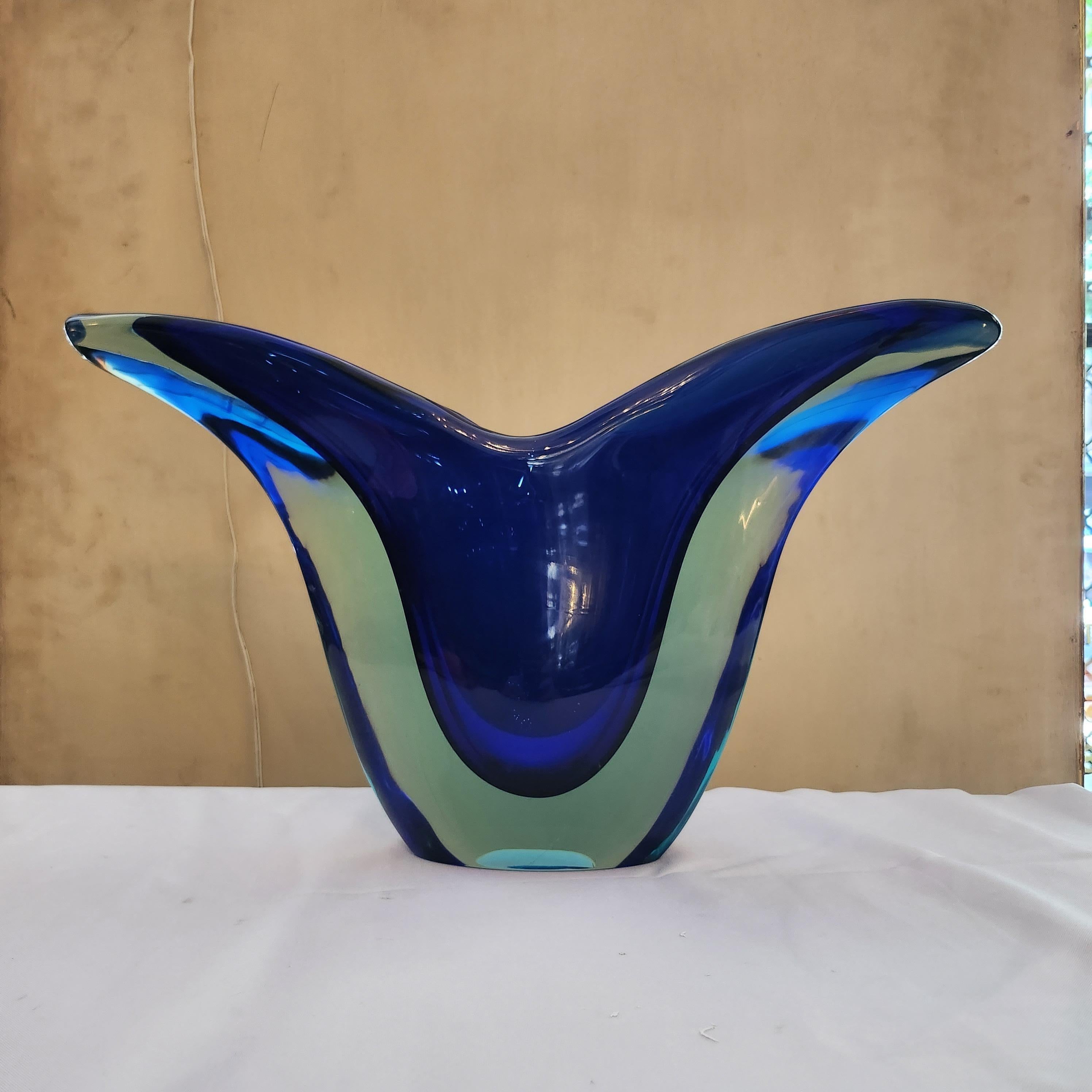 This Flavio Poli sommerso glass vase, dating back to the vibrant era of the 1970s, exudes an air of timeless elegance and sophistication. Crafted in a striking V form, it seamlessly blends shades of deep blue with transparent glass, creating a