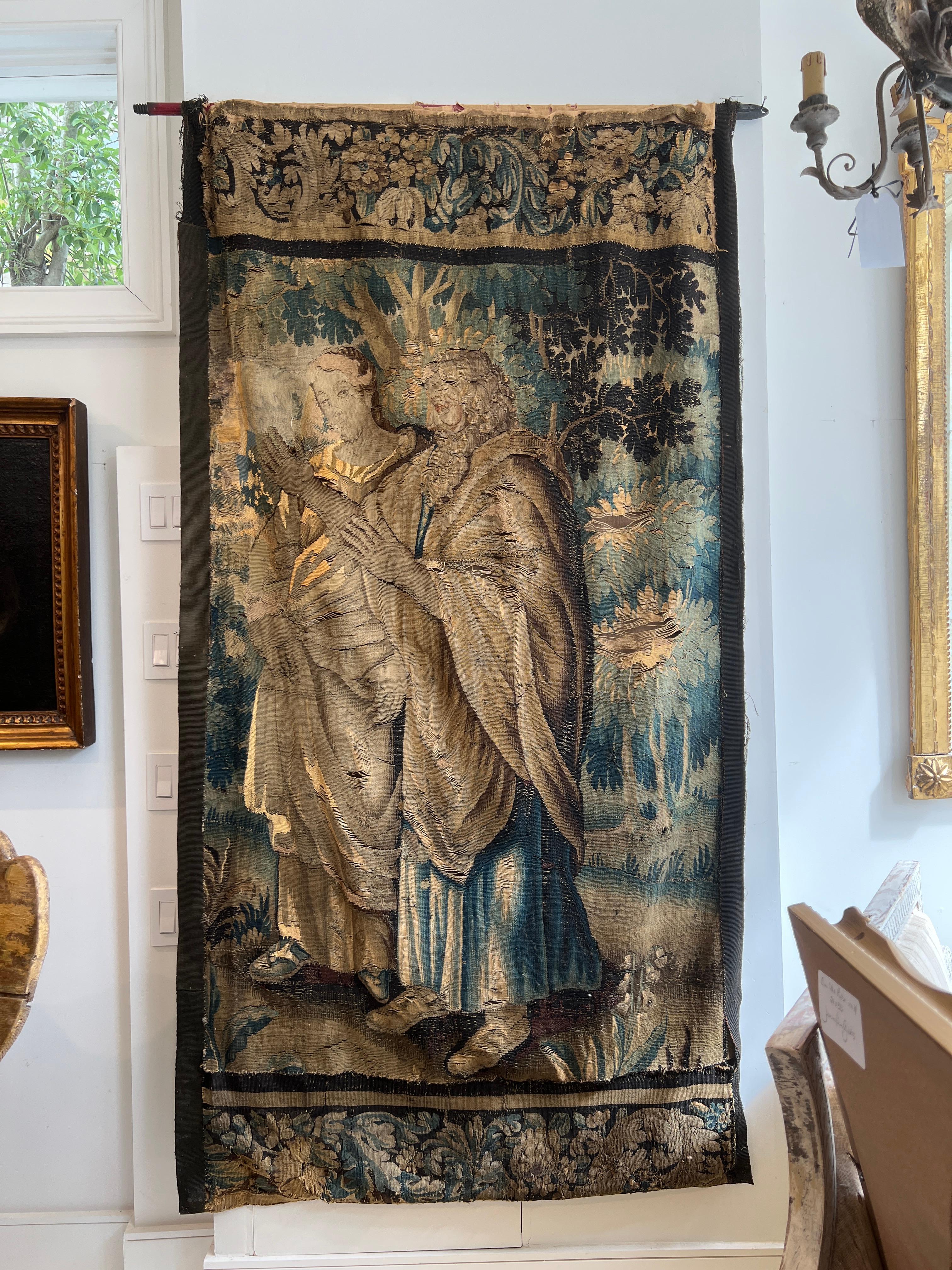 This Flemish 18th Century tapestry features a man and woman in a landscape setting with a floral border above and below. Frayed edges and losses in fabric indicate age. Unusual size indicates it could be a portion of an originally larger work.