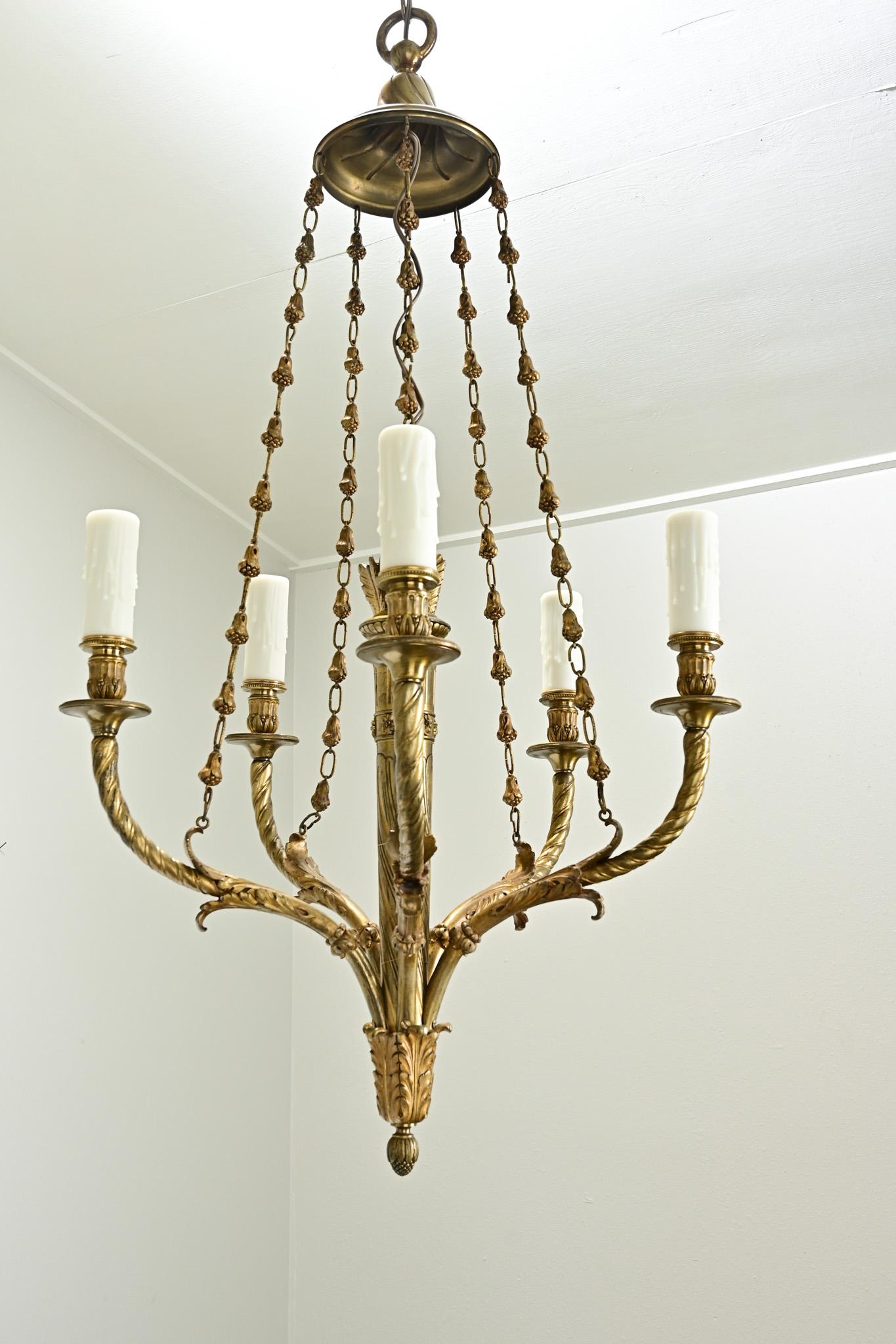 An elegant 5-light Louis XVI style chandelier made in Belgium. This heavy brass light fixture is centered with a brass quiver of arrows and has five long arms ending in candle cups with faux wax candle covers. A decorative chain connects the