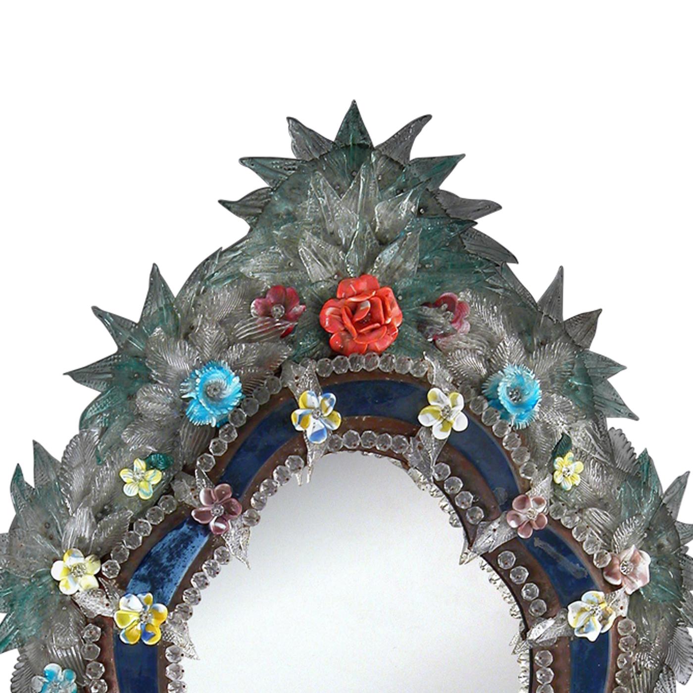 This elegant mirror has an antique finish and an exquisite blue frame in Murano glass adorned at the top and bottom by hand-made vivid green leaves and multicolored flowers.