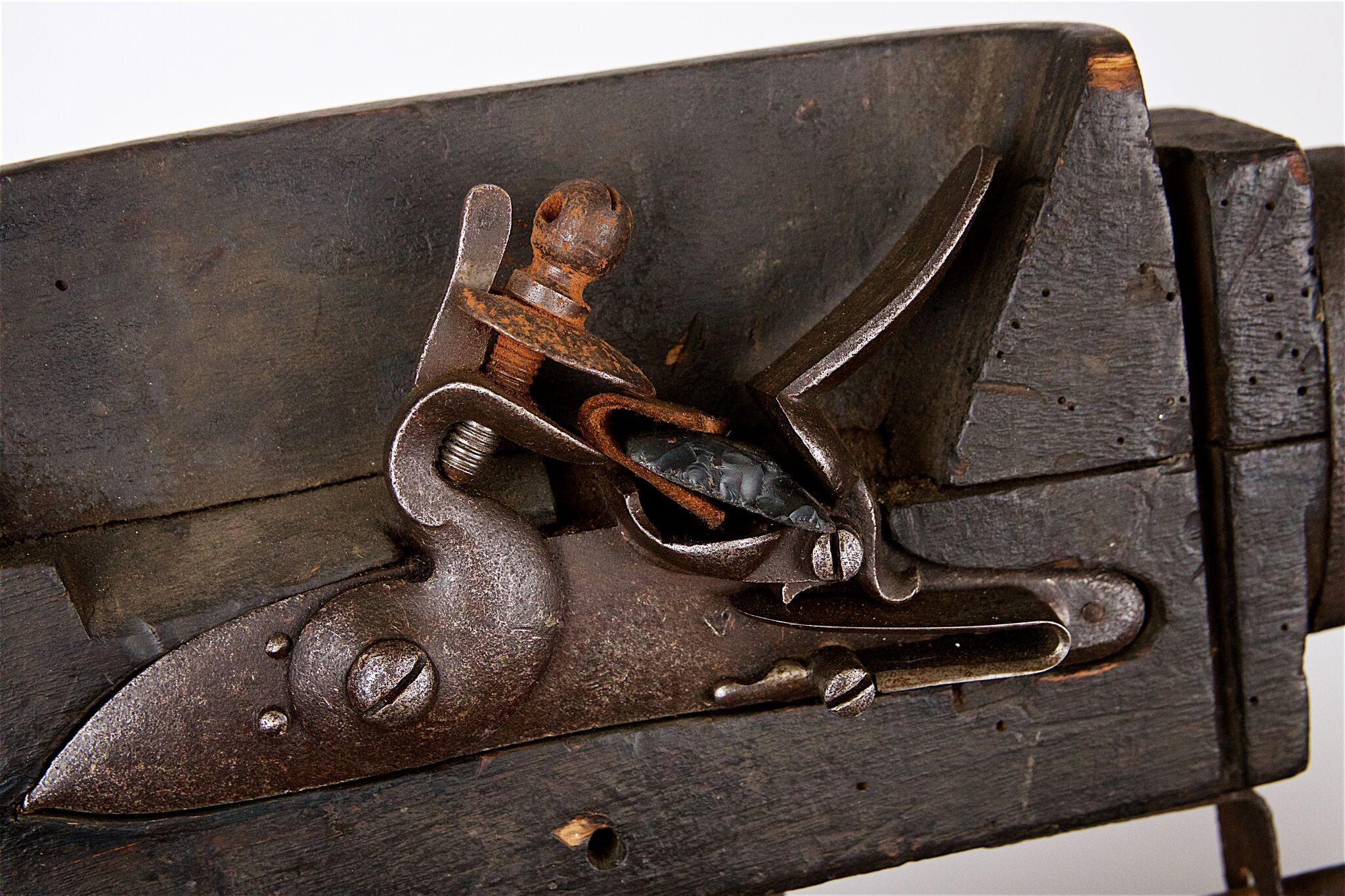 Flintlock alarm gun with simple flared blunderbuss style barrel and flint-fired musket lock mounted to an unusual wooden casing. The gun has an iron pintle underneath with a sliding trigger bar which allows the forward motion of a tripwire to pull