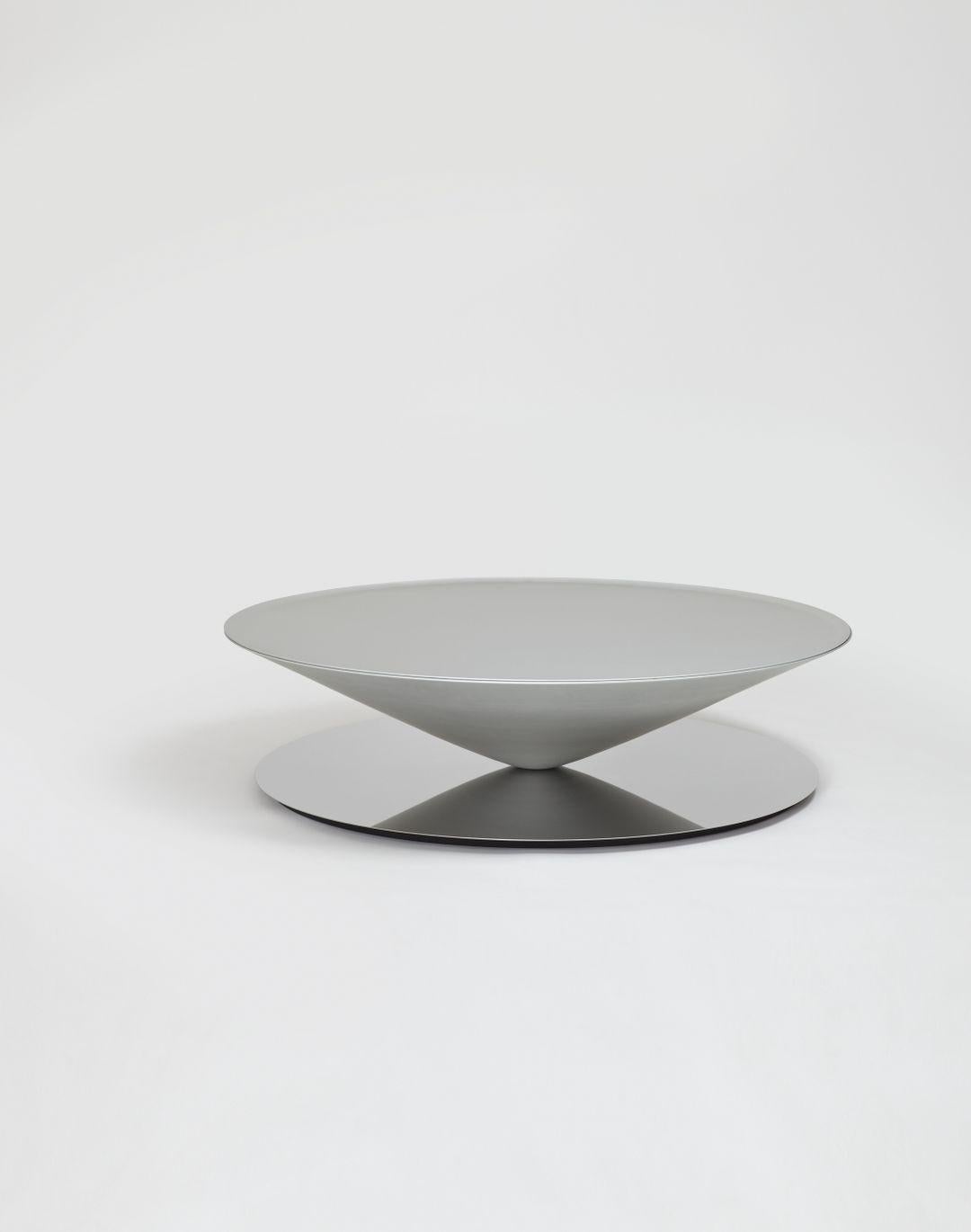 Float est une table basse sculpturale qui défie les sens et la perception. Un cône massif en métal flotte apparemment au-dessus d'une base en acier poli miroir. Le design géométrique est adouci par des détails raffinés tels que la lèvre biseautée