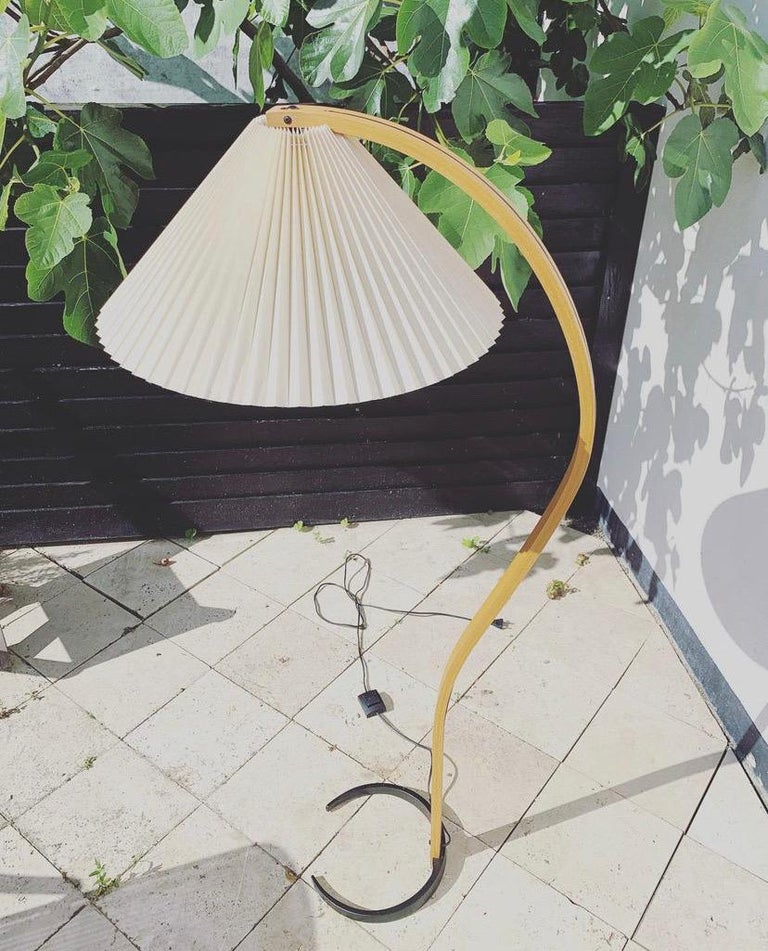 This beautiful and iconic floorlamp was designed by Danish Mads Caprani in the 1970s. This vintage lamp features a sculptural bent beech stand with an elegant curve, a cast iron crescent shaped base, and original pleated linen shade. The original