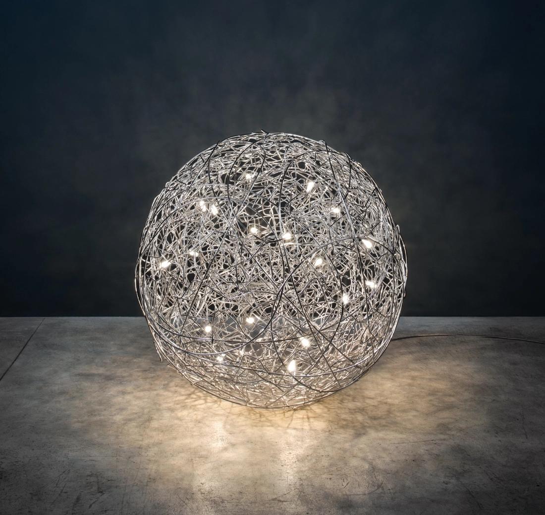 Fil de Fer, disegnata da Enzo Catellani, è una lampada da terra realizzata con fili di alluminio sagomati e intrecciati, illuminati dall'interno. La sua sfera compatta emette una luce soffusa e intima, perfetta per creare angoli accoglienti e