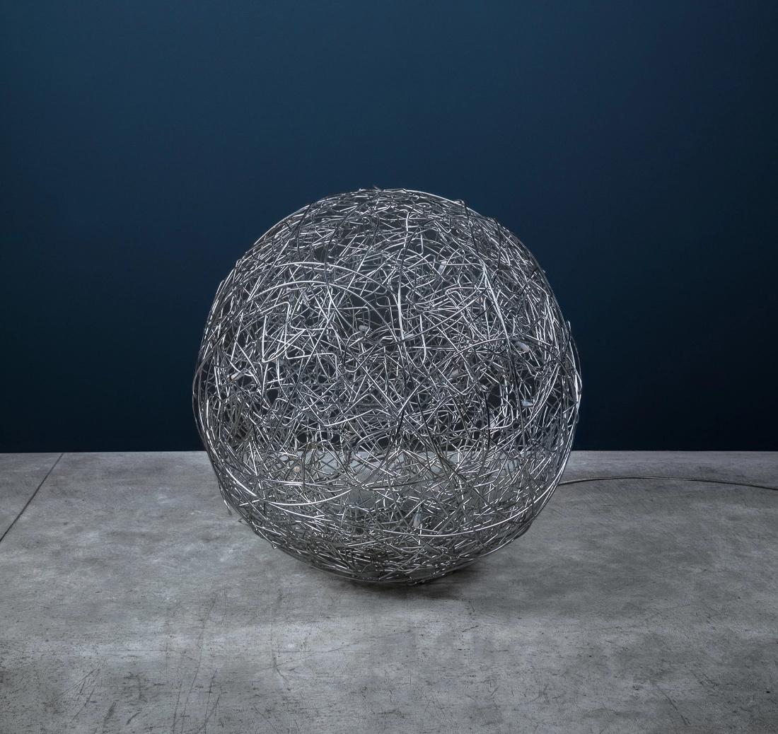 La Fil de Fer, diseñada por Enzo Catellani, es una lámpara de pie de autor fabricada con alambre de aluminio trenzado e iluminada desde dentro. Su estructura mínima y su suave resplandor interno crean una presencia mágica y atmosférica en cualquier