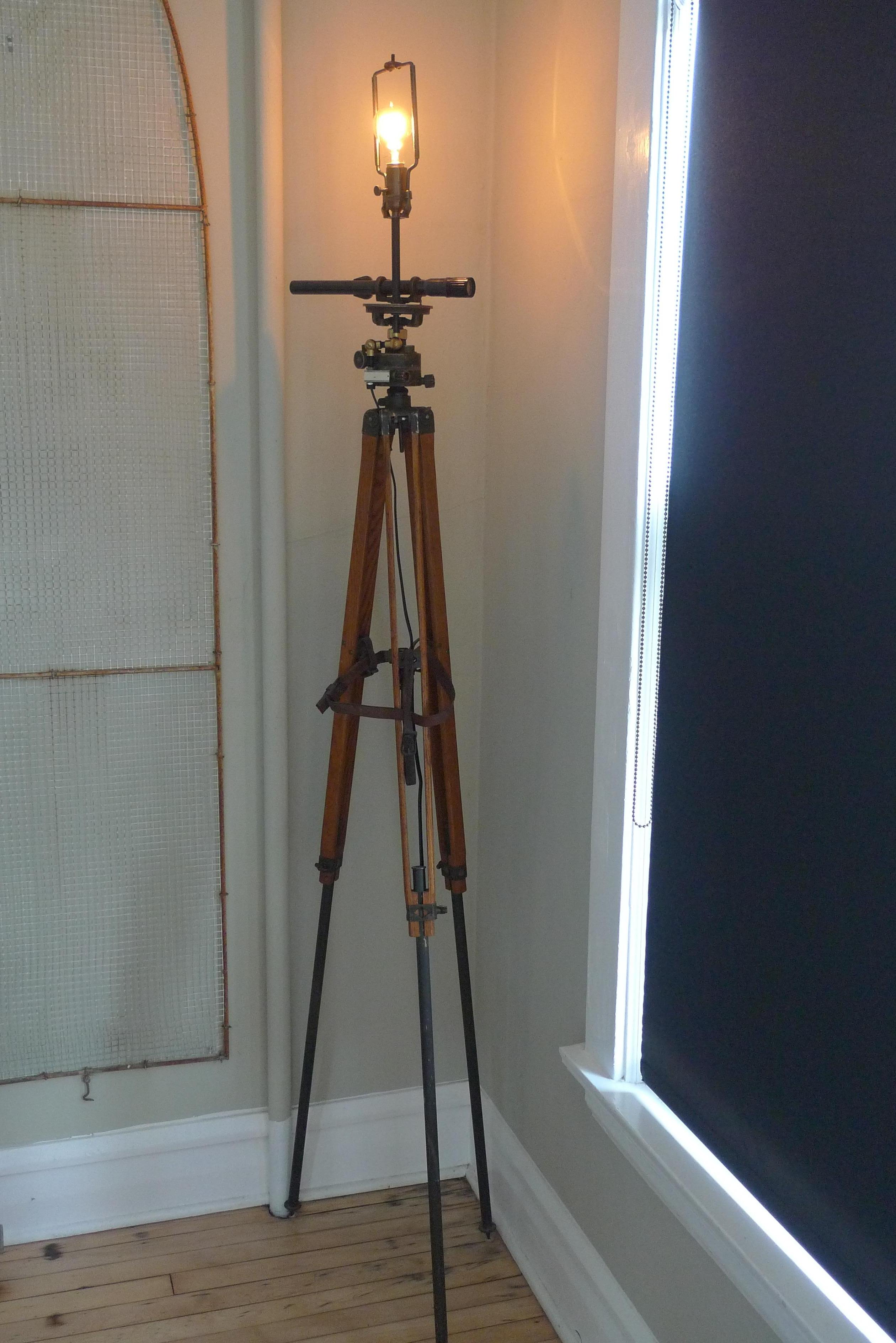 Floor lamp made from U.S. Army surveyor tripod (circa 1940s) with scope, oak legs and leather strapping. Shade not included. Height is adjustable from 48 to 70 inches. Leather strapping helps anchor tripod legs. Light has 3-way socket, brass harp