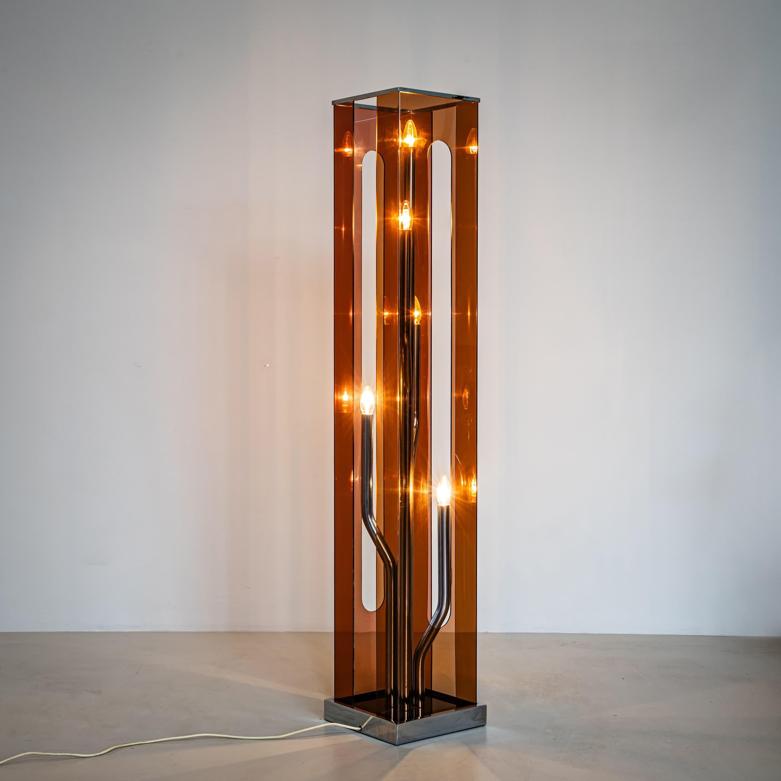 Lámpara de pie con pantalla de plexiglás violeta con recortes alargados, colocada sobre una base cuadrada de metal. En su interior, cinco varillas metálicas dobladas terminan cada una en un enchufe. Una elegante fuente de luz indirecta con una