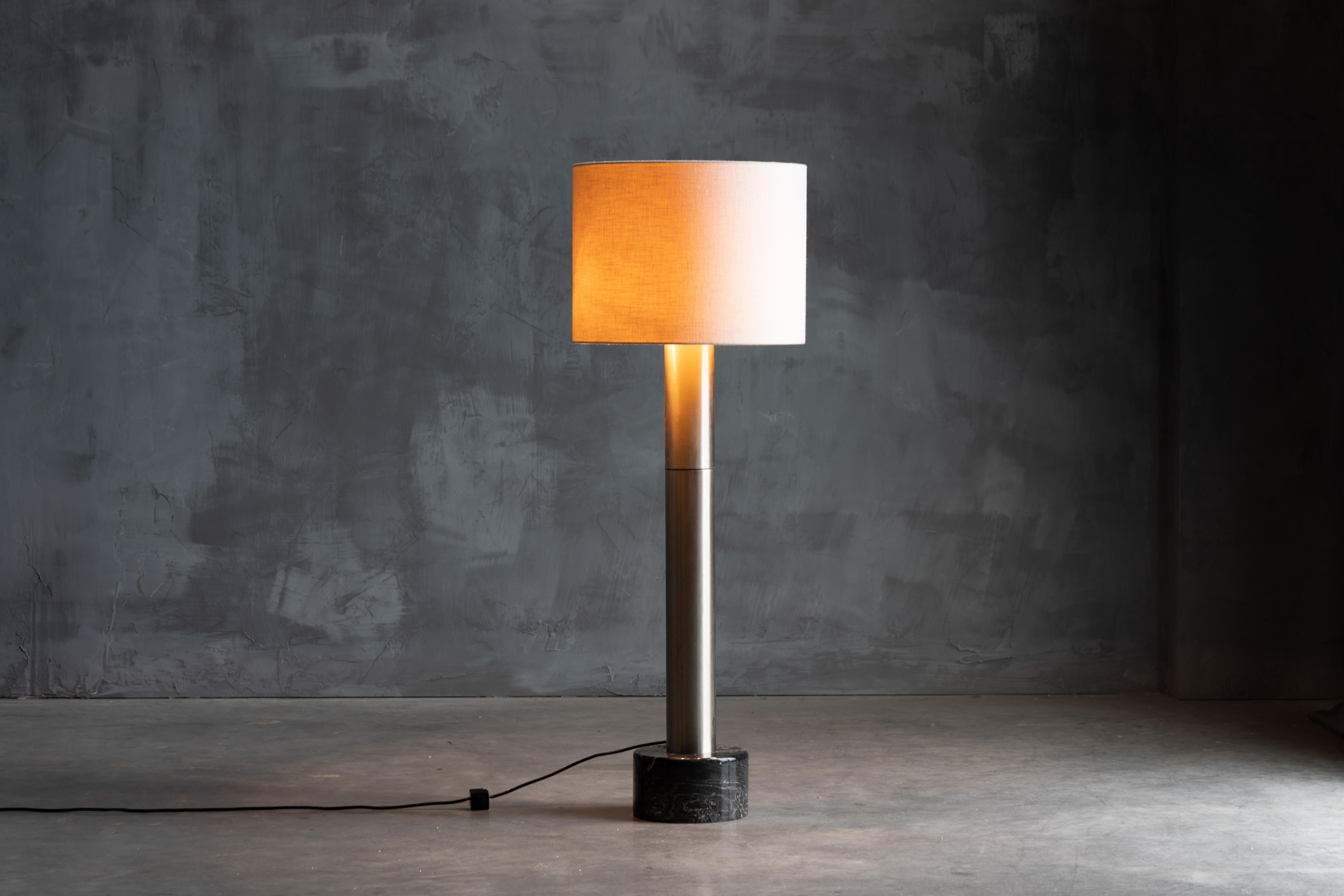 Brutalist metal floor lamp, a fusion of industrial edge and refined design. Its bold silhouette stands tall atop a sleek black marble round base. Illuminate your surroundings with this floor lamp, as it commands attention while casting a warm,
