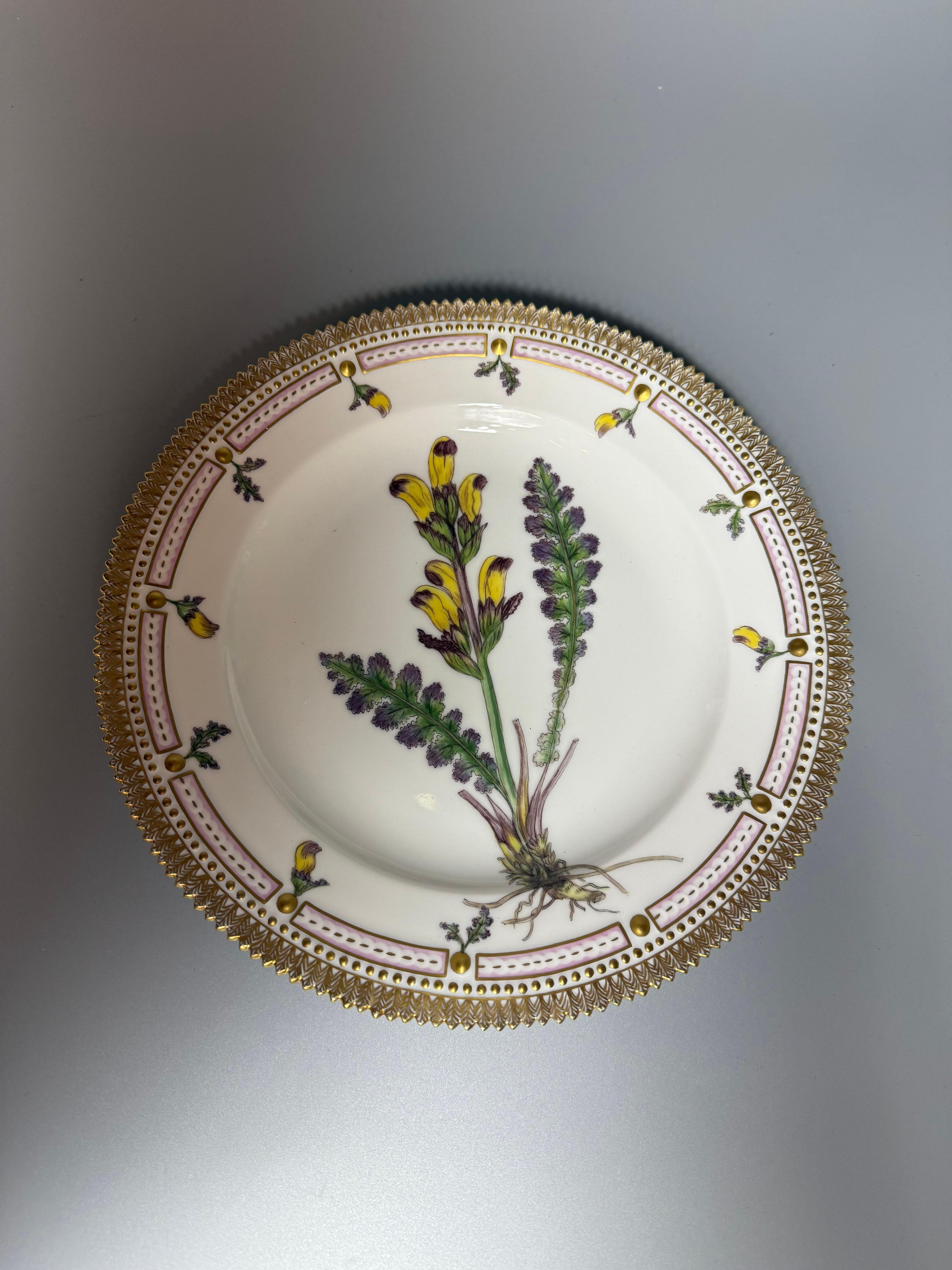 L'image montre une assiette à dîner Flora Danica de Royal Copenhagen. 
Cette collection de porcelaine est réputée pour ses illustrations botaniques exquises peintes à la main, inspirées de l'encyclopédie botanique des XVIIIe et XIXe siècles, Icones