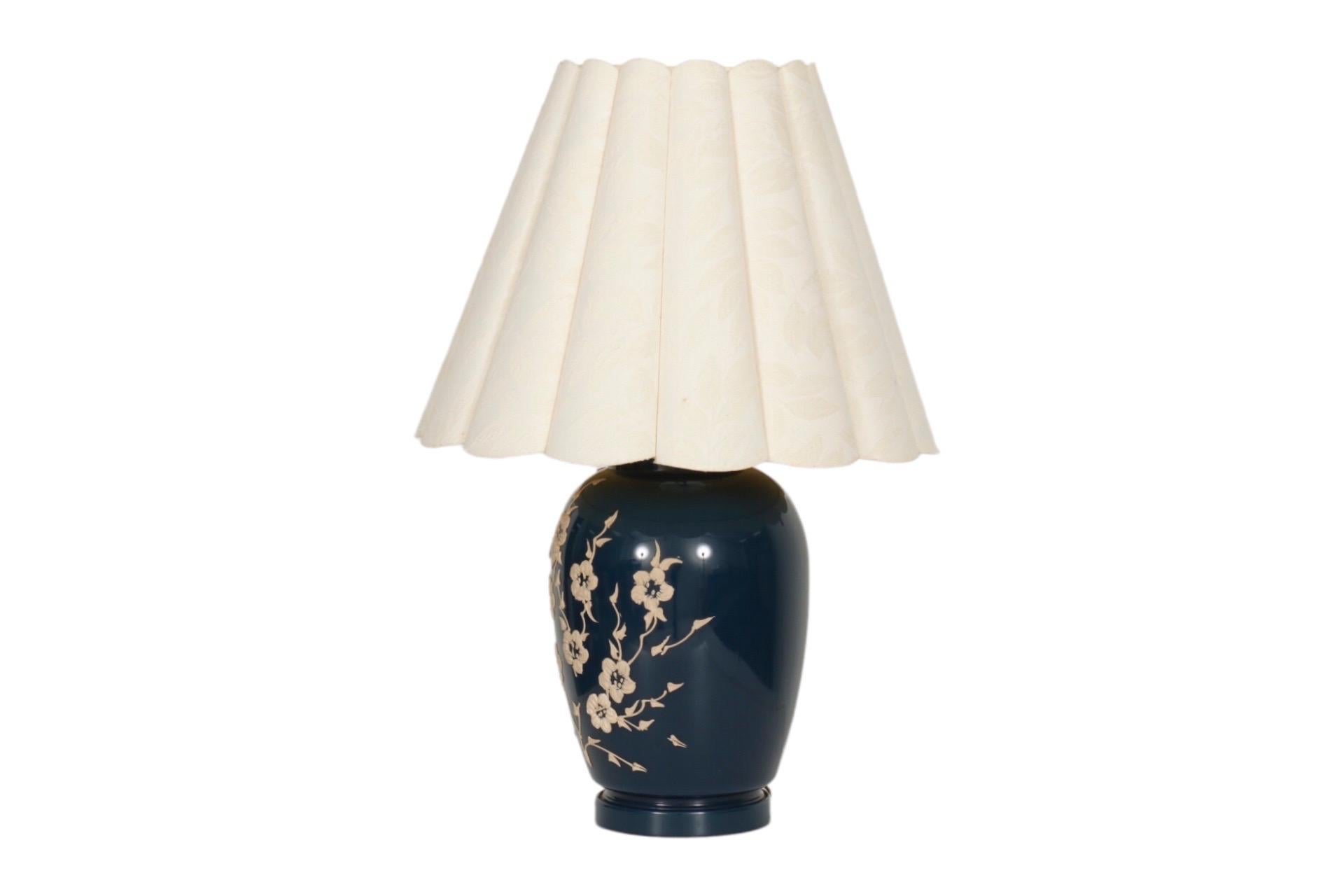 A navy blue ceramic table lamp. The ginger jar shaped vase is decorated with elegantly applied flowers in tan. Topped with a rounded flute lampshade in cream. Measures 19”H to the top of the socket, 26.75”H to the top of the finial. Shade measures