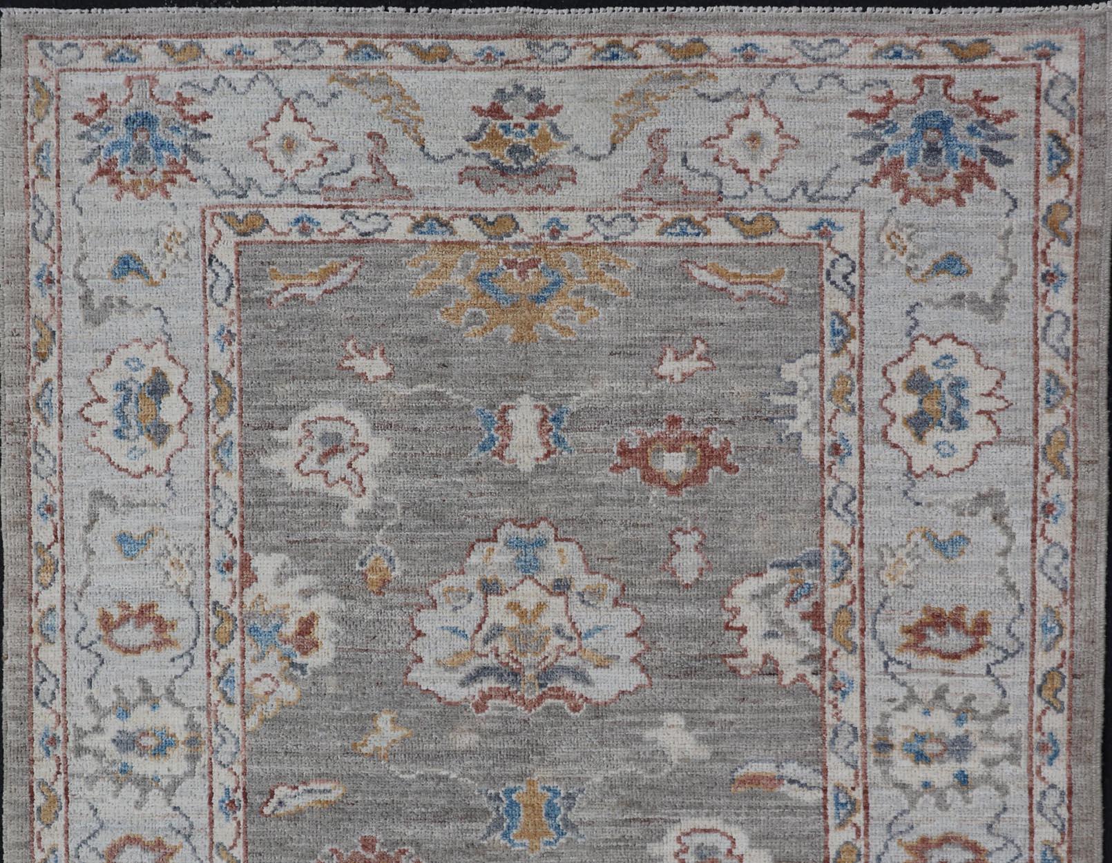 Oushak moderne présentant un motif floral en forme d'arabesque sur toute la surface. L'arrière-plan est rendu dans un gris clair, tandis que la bordure est d'un bleu étain clair. Le motif présente des couleurs ivoire, or, orange rouille et brun.