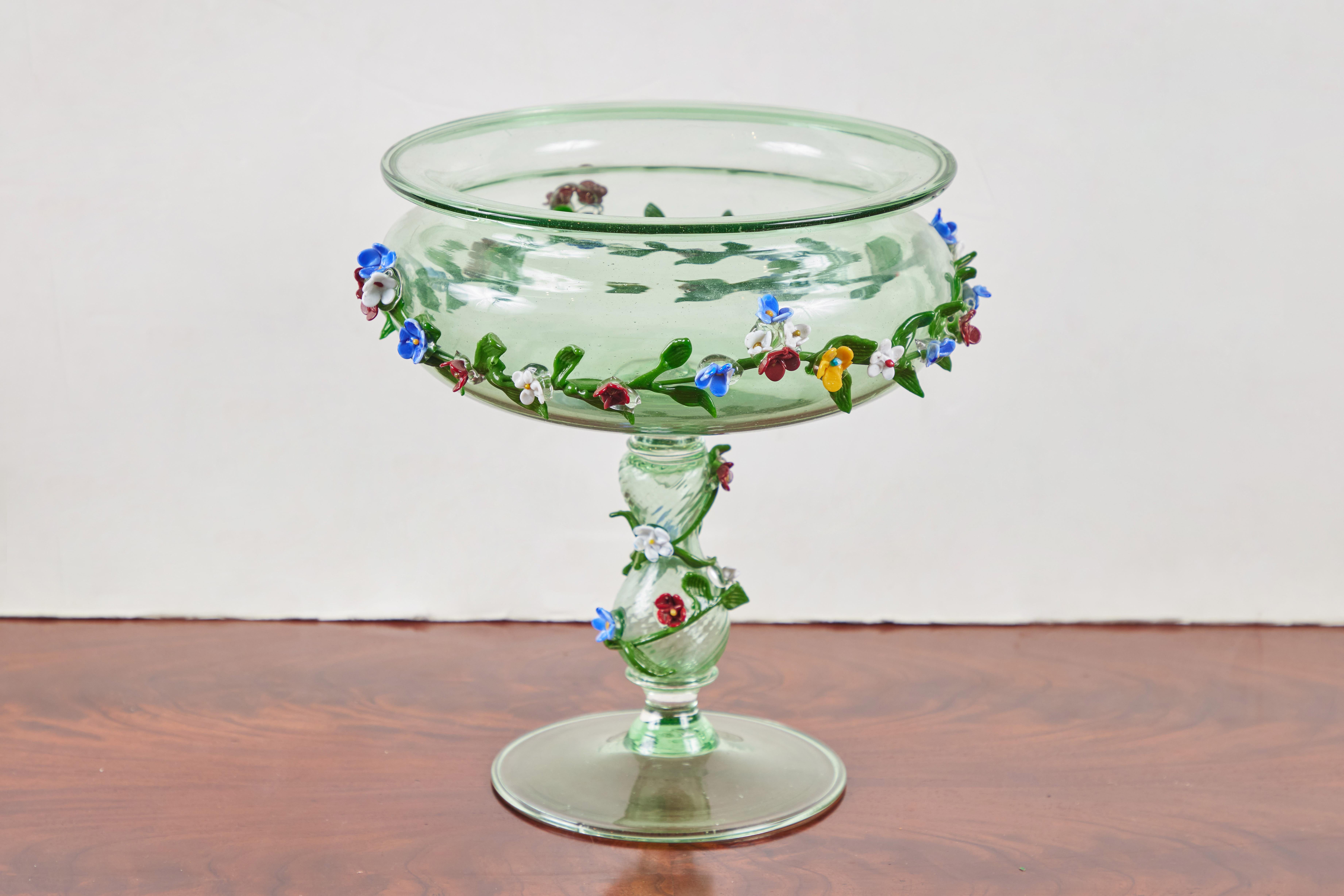 Hübsche, um 1920, mundgeblasene Tazza aus Murano-Glas in frischem Frühlingsgrün. Die lippenförmige Schale ist mit einer applizierten Girlande aus individuell geformten Blüten und Blättern in verschiedenen Farben geschmückt, die sich am Stiel