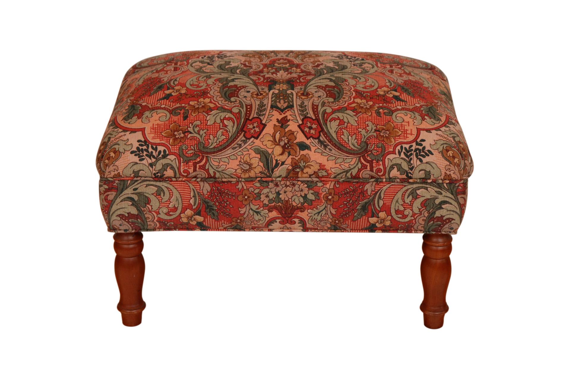 An English style ottoman. Upholstered in a rich Ishana style floral printed cotton with bunches of acanthus foliage repeated in red, sage, mocha and olive on a blush ground. Stands on turned wooden legs finished in blunt arrow feet.