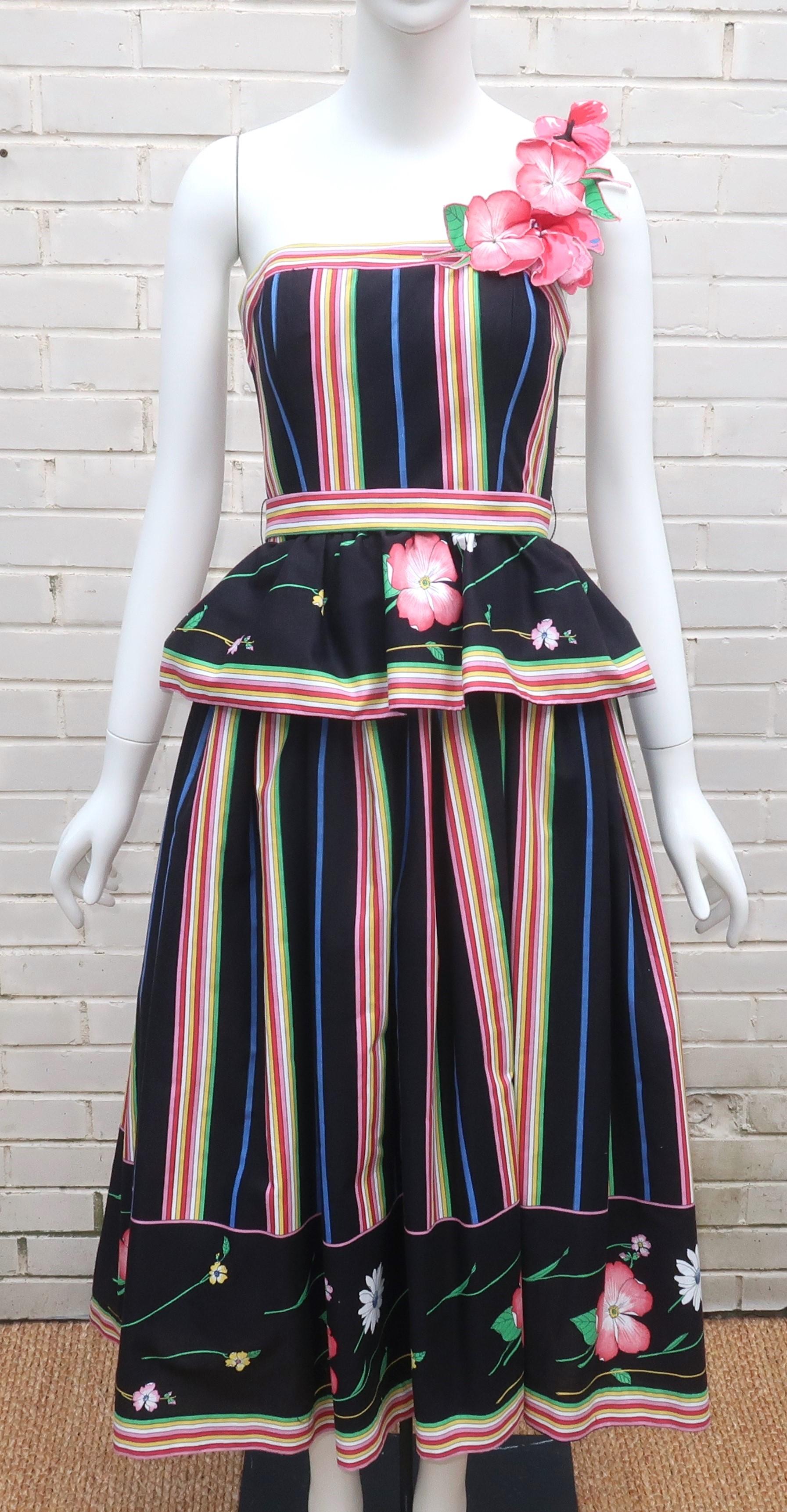 1970's polished cotton sundress in a floral and stripe print with a mix of cheerful colors including pink, red, yellow, green and blue on a black background.  The one shouldered design has a peplum waist and full skirt lined with a netted crinoline.