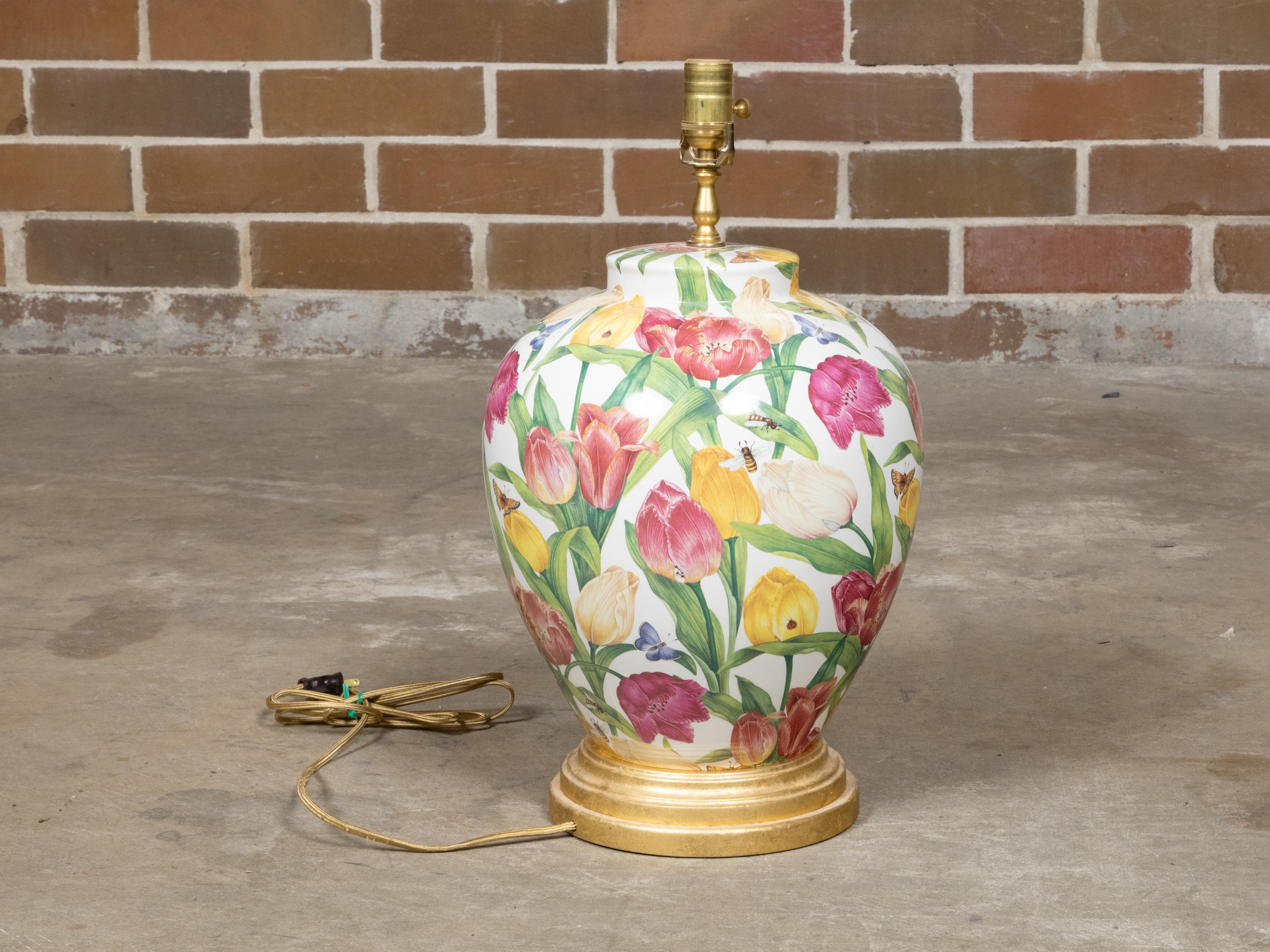 A colorful floral porcelain table lamp with single socket and circular giltwood base. This charming porcelain table lamp captivates with its vibrant floral décor, showcasing a delightful array of tulips, bees, and butterflies, all rendered in vivid