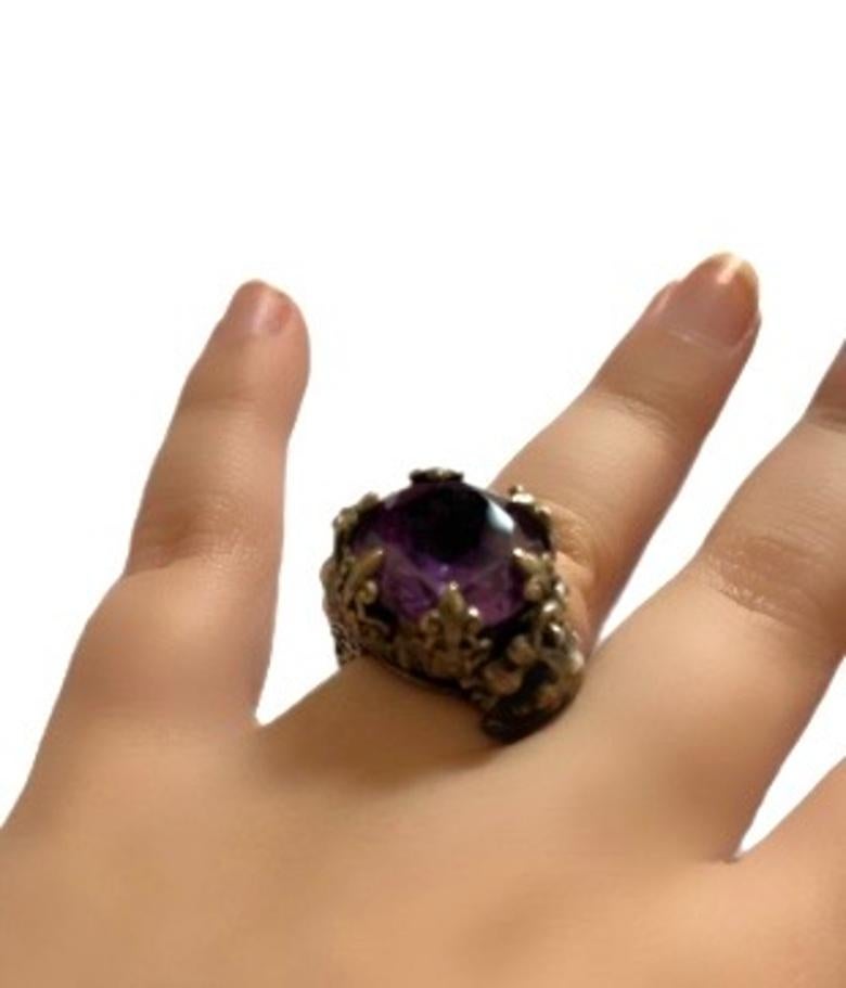 Aggiungi un tocco di fascino vintage alla tua collezione di gioielli con questo squisito anello regolabile. È incastonata in un'intricata lavorazione floreale in metallo in stile antico, con gigli che circondano e sostengono la pietra centrale. Il